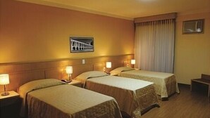 Habitación de hotel con dos camas, cabecero, mesita de noche, lámpara, ventana con cortinas e imagen colgada en la pared.