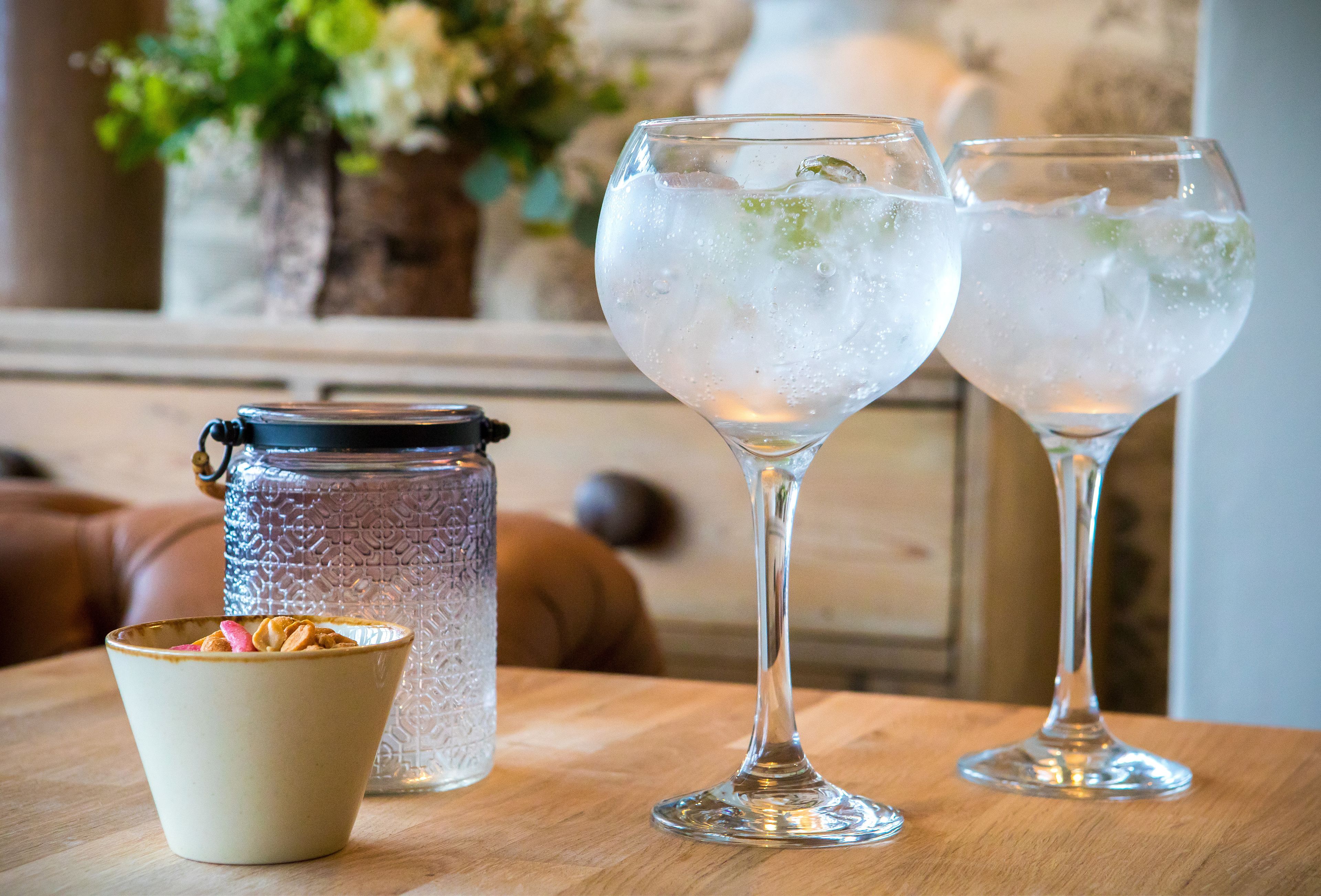 Two glasses of iced beverage on a wooden table, a ceramic bowl with nuts, and a glass jar with a metal lid.