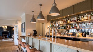 A bar area with a wooden counter, multiple bar stools, and a variety of liquor bottles. - Beaulieu Hotel (Brockenhurst)