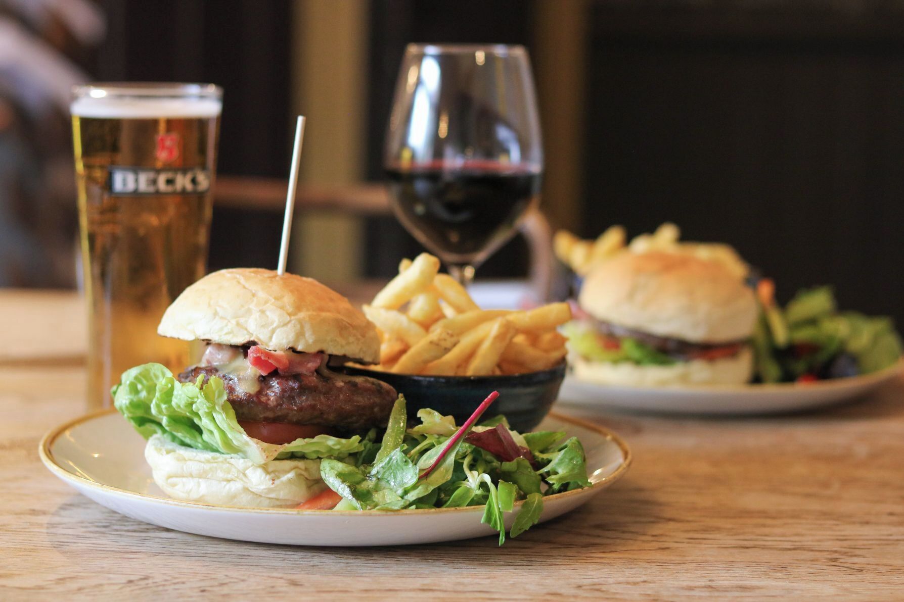 Un plato con una hamburguesa, papas fritas y una ensalada, acompañado de una jarra de cerveza y una copa de vino tinto.