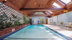 Una piscina interior con techo de madera, pared de ladrillo con plantas y zona poco profunda.