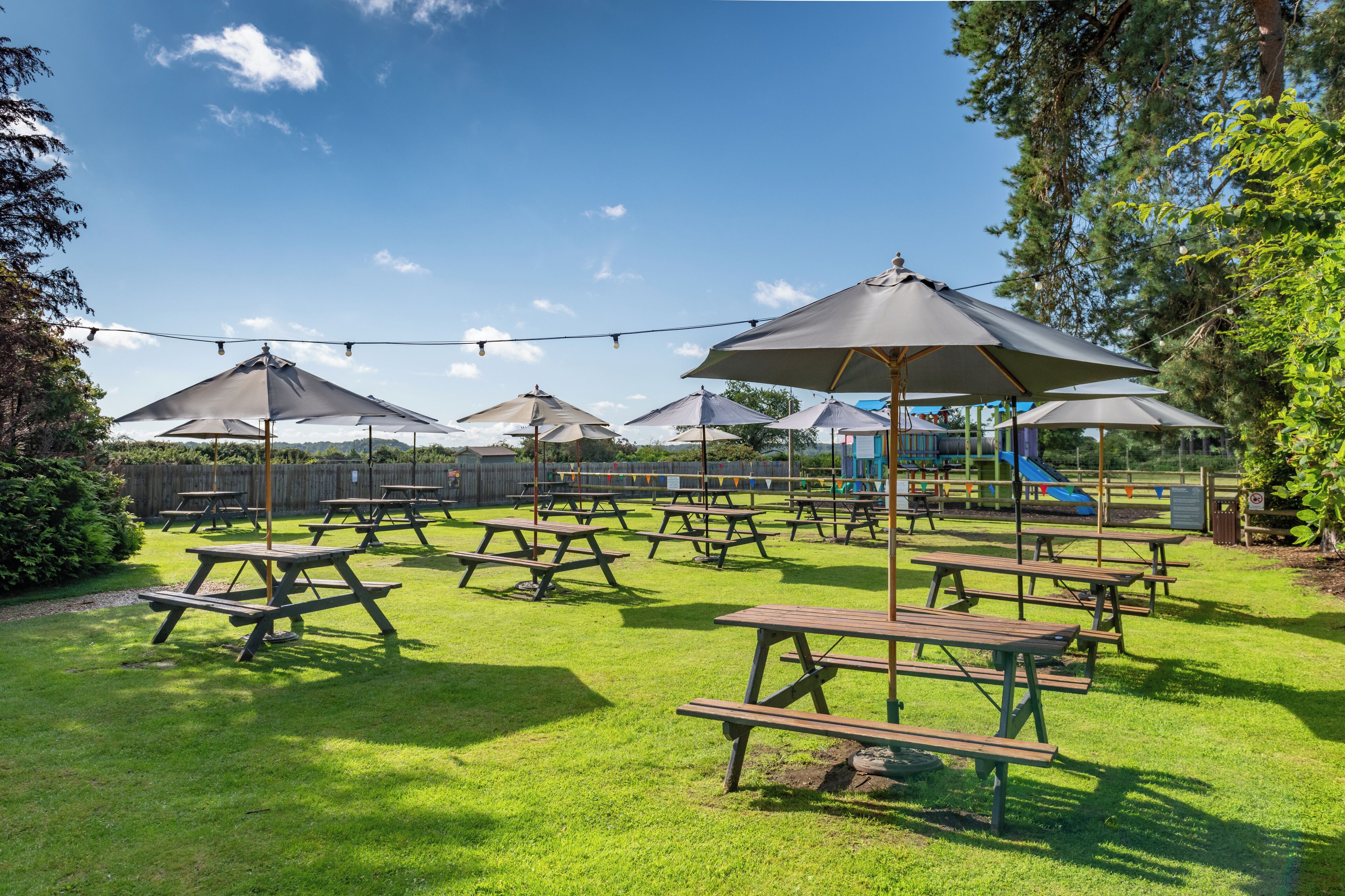 An outdoor area with picnic tables and umbrellas, surrounded by trees and a playground.