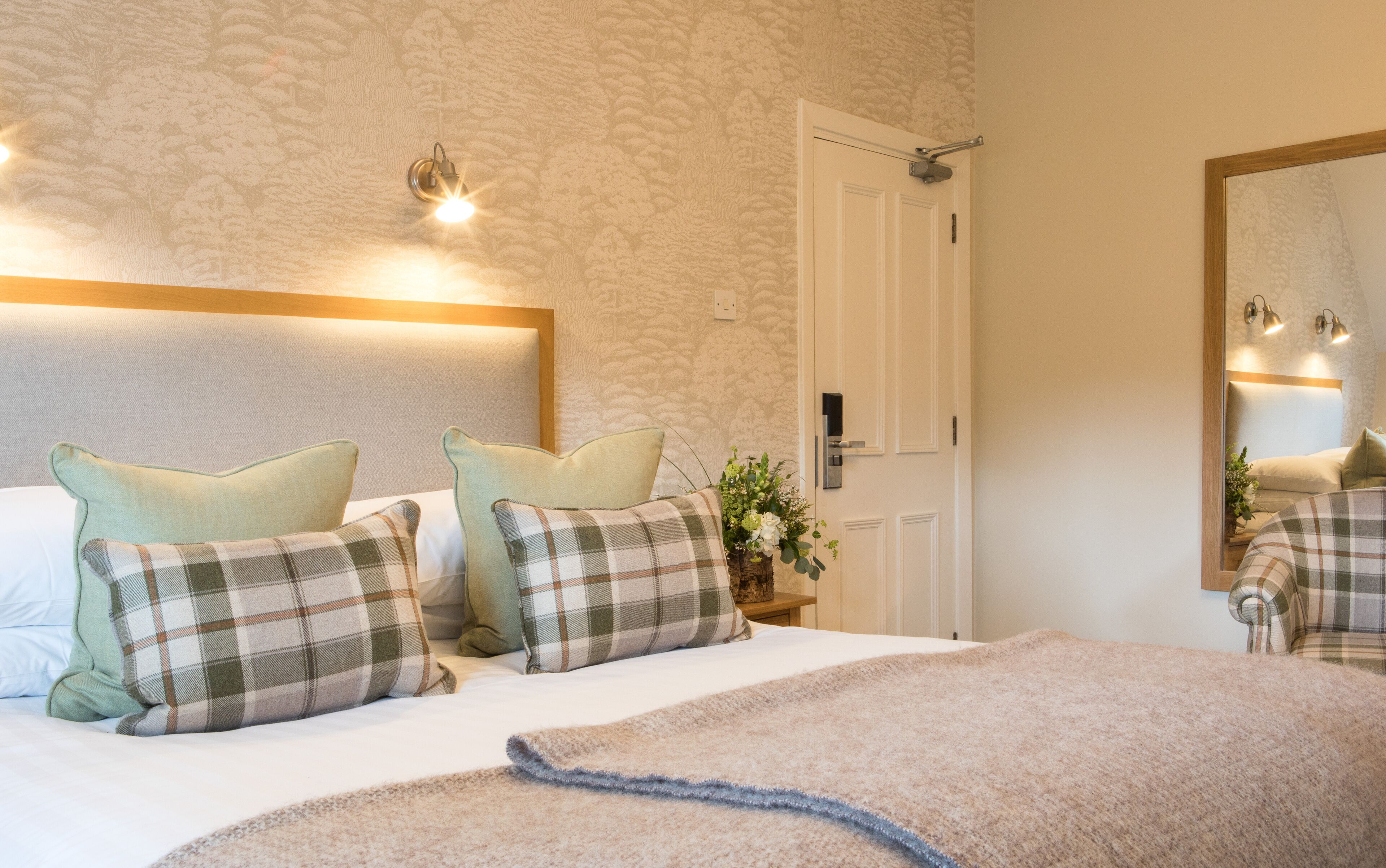 A neatly made bed with plaid pillows, a bedside table with a vase of flowers, and a wall-mounted mirror.