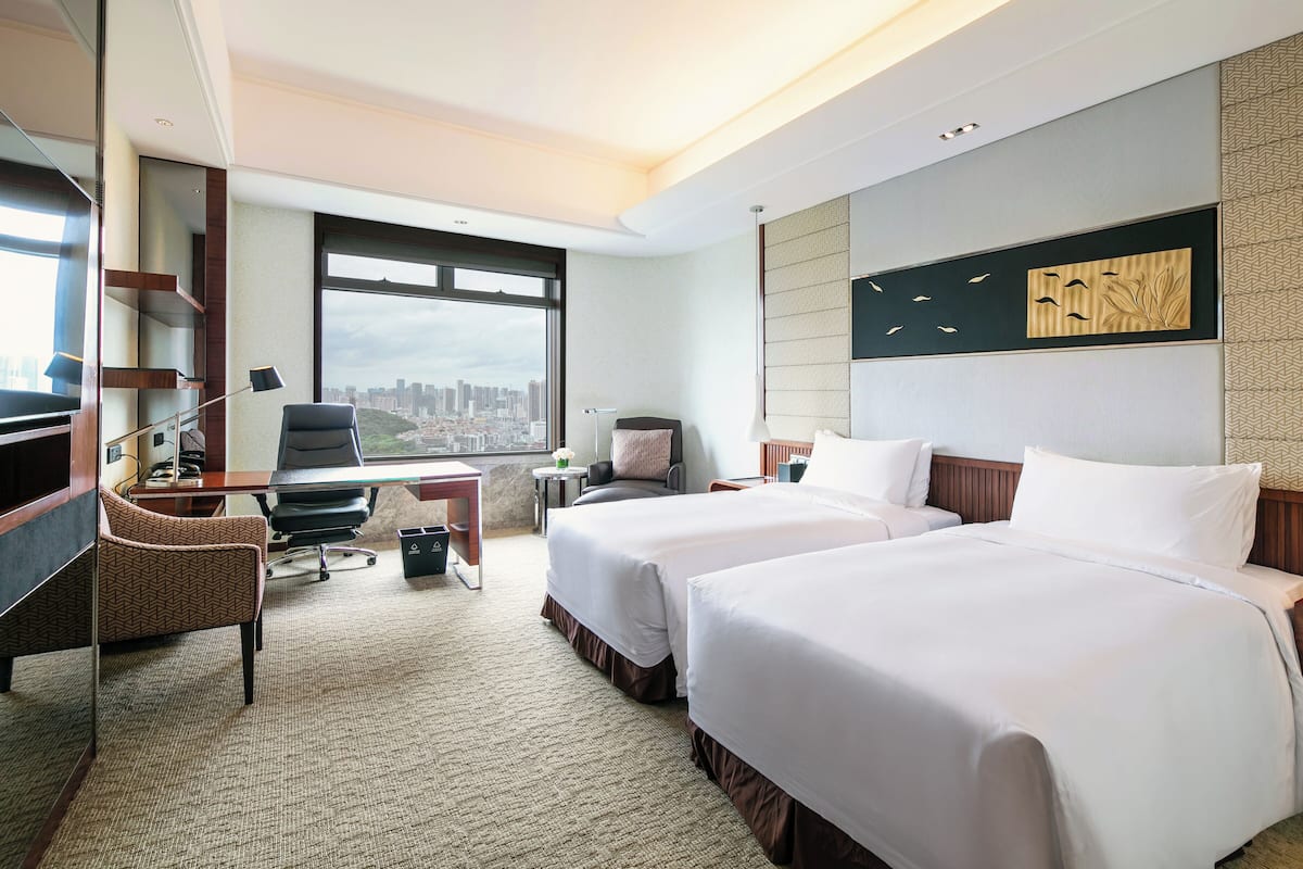 A hotel room with two beds, a desk, a chair, a small table, and a large window with a city view.