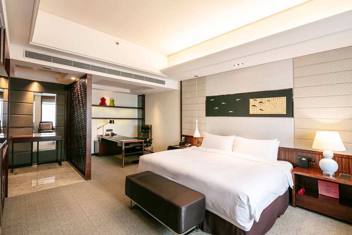 A hotel room with a large bed, a desk, a chair, a bench, and a nightstand with a lamp.