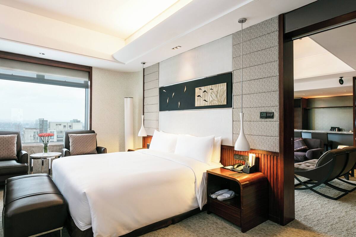 A hotel room with a large bed, a desk, a chair, and a sofa. There is a window with a view of buildings.
