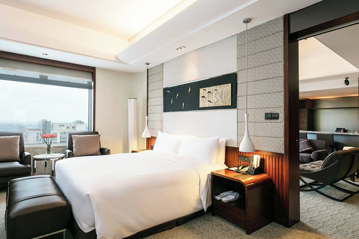 A hotel room with a large bed, a desk, a chair, and a sofa. There is a window with a view of buildings.