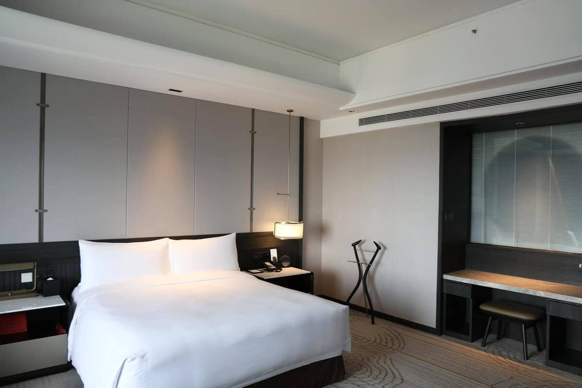A hotel room with a large bed, bedside tables, a desk, and a window with blinds.