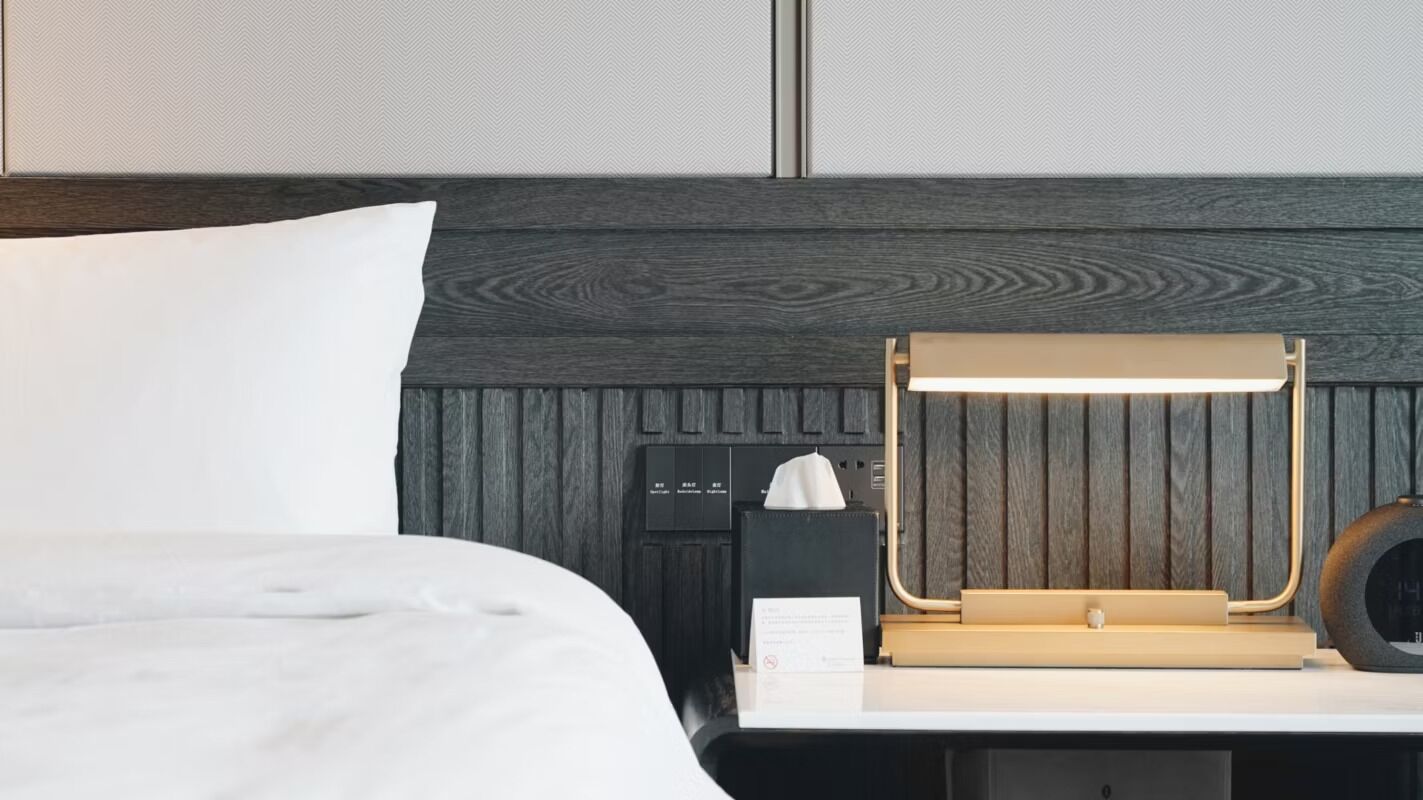 A hotel room with a bed, a bedside table with a lamp, a nightstand, and a wall panel.