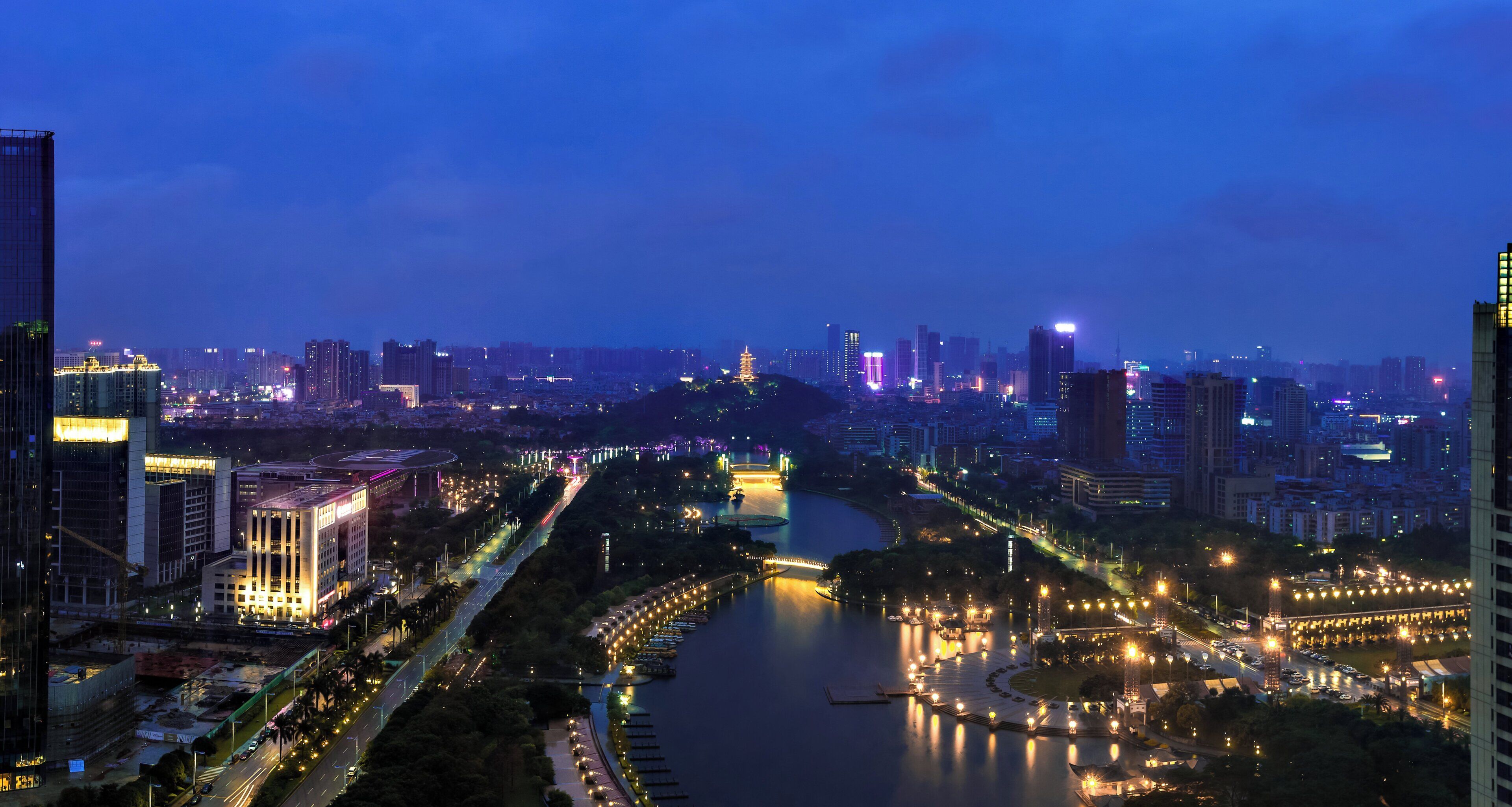 Paisagem urbana ao entardecer com edifícios iluminados, um rio e uma ponte.