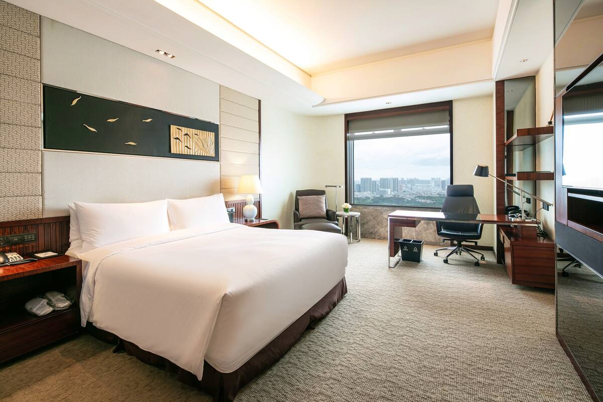 A hotel room with a large bed, a desk, a chair, a TV, and a view of the city.