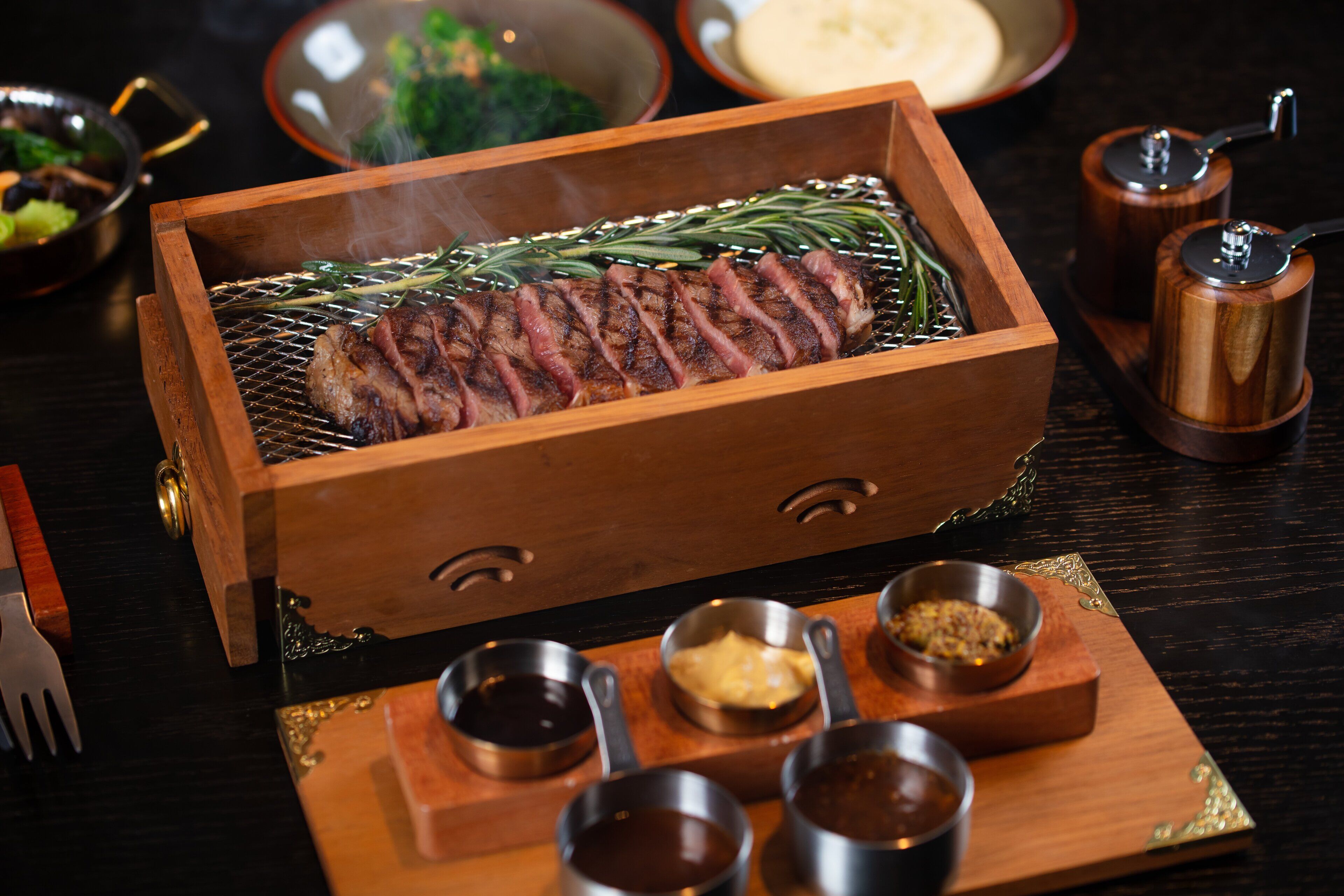 A wooden grill with a steak, surrounded by condiments and a fork on a wooden tray.
