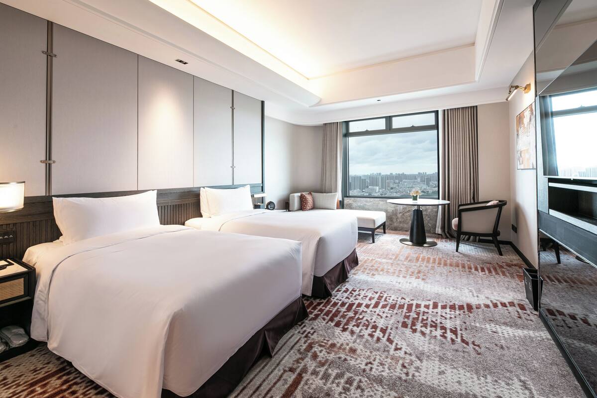 A hotel room with two beds, a desk, a chair, a TV, and a city view.