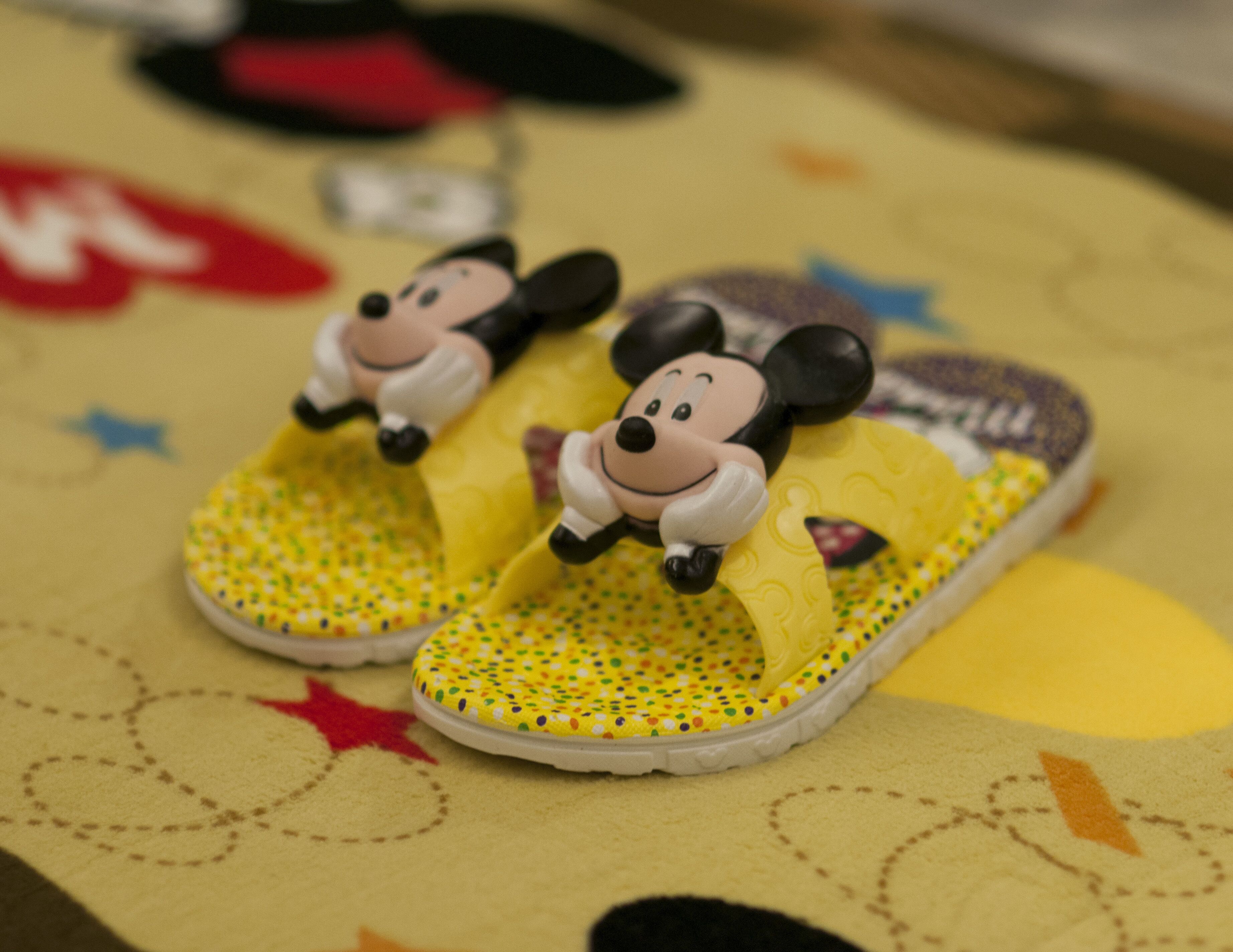 Une paire de chaussons à l’effigie de Mickey Mouse, posée sur un tapis jaune orné de motifs en forme d’étoiles.