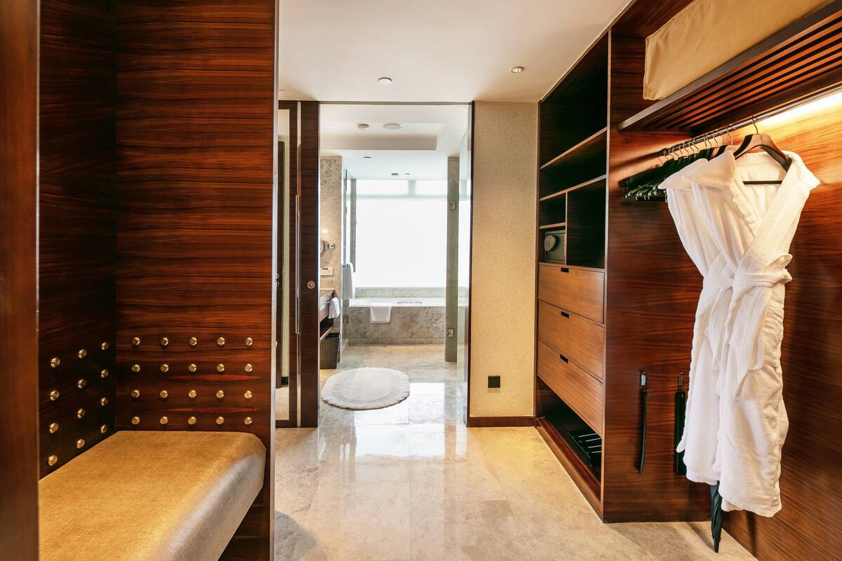 A walk-in closet with wooden paneling, a robe hanging on a rack, and a view into a bathroom with a bathtub and a toilet.