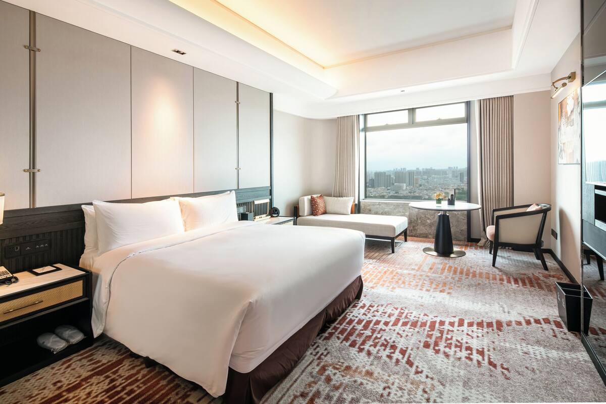 A hotel room with a large bed, a desk, a chair, a small table, and a view of the city.