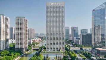 A high-rise building with a grid of windows, surrounded by other tall buildings, a river, and greenery.
