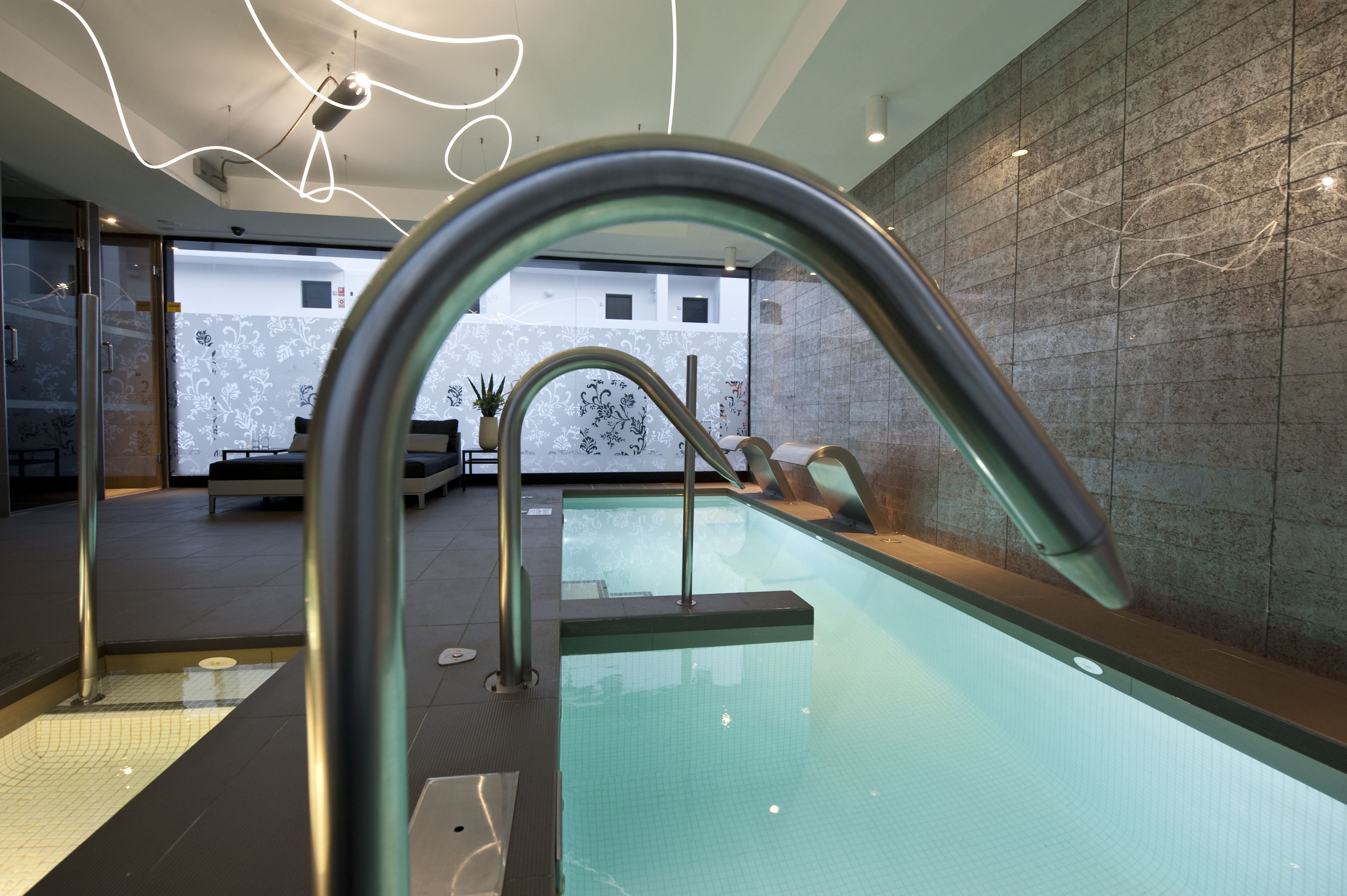Una piscina interior moderna con agua cristalina, barandillas metálicas y una pared de azulejos con un diseño geométrico.