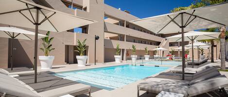 A poolside area with lounge chairs and umbrellas, a swimming pool, and a building with multiple balconies.