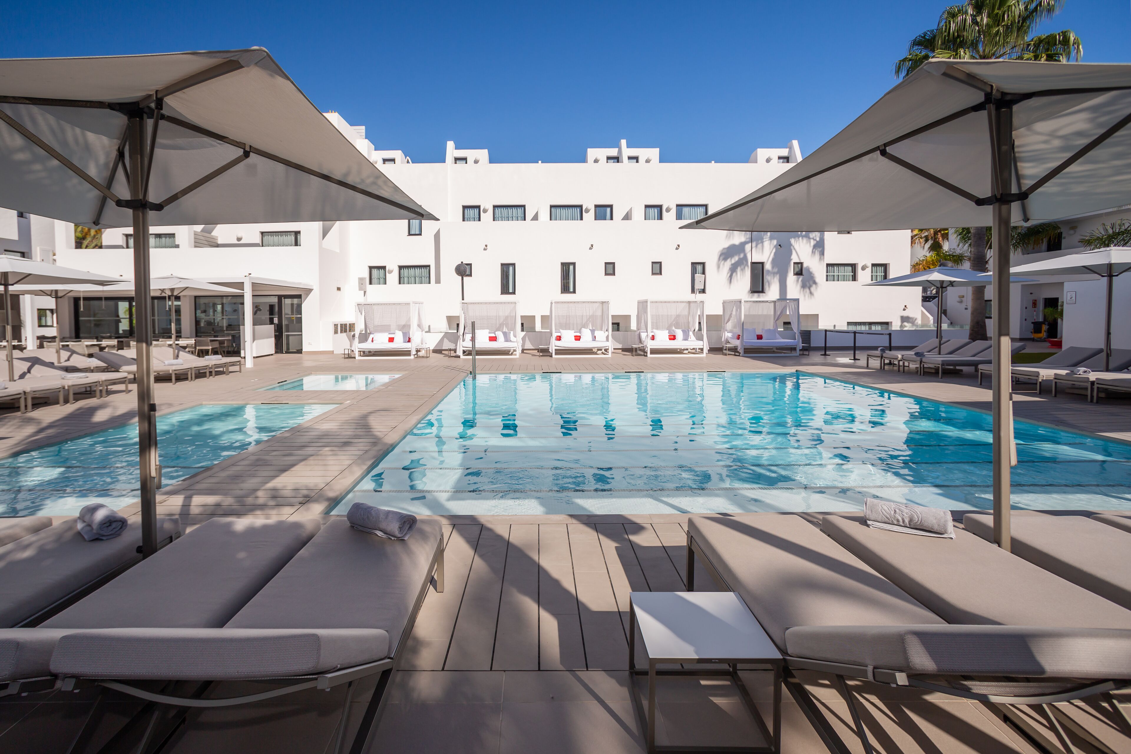 A pool area with lounge chairs and umbrellas, a clear blue sky, and a modern building in the background.