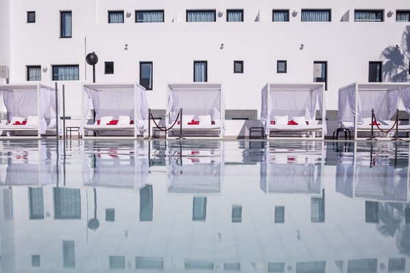 Un hôtel doté de nombreuses chambres, chacune comprenant des lits avec du linge de lit blanc et des touches de couleur rouge, et disposées en rangée le long d’un bassin aux reflets scintillants.