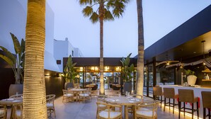 An outdoor dining area with wicker tables and chairs, palm trees, and a modern building in the background. - Migjorn Ibiza Suites & Spa (Sant Josep de sa Talaia)