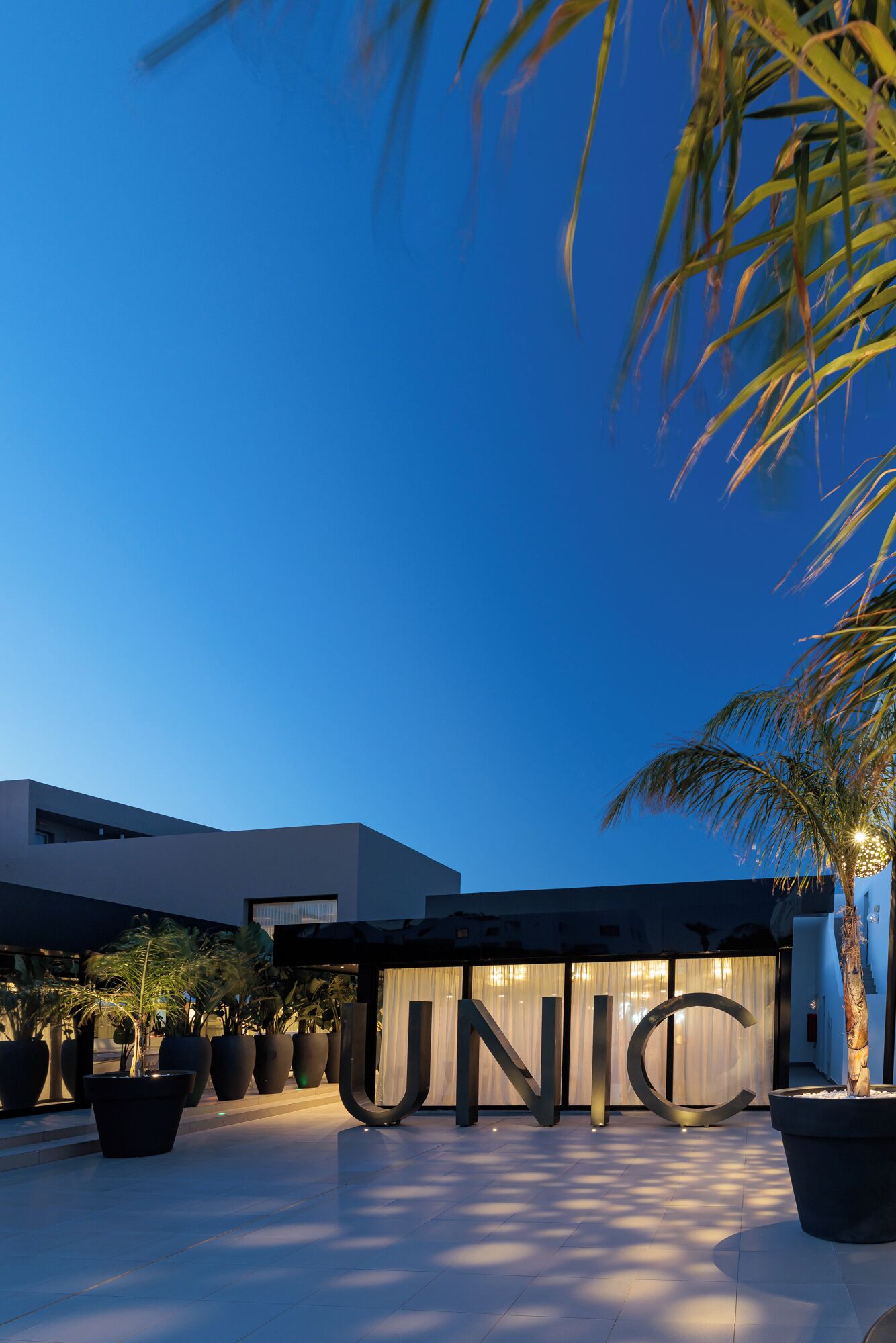 Ein modernes Gebäude mit einer großen, beleuchteten 'UNIC'-Beschriftung, Pflanzkübeln und blauem Himmel.