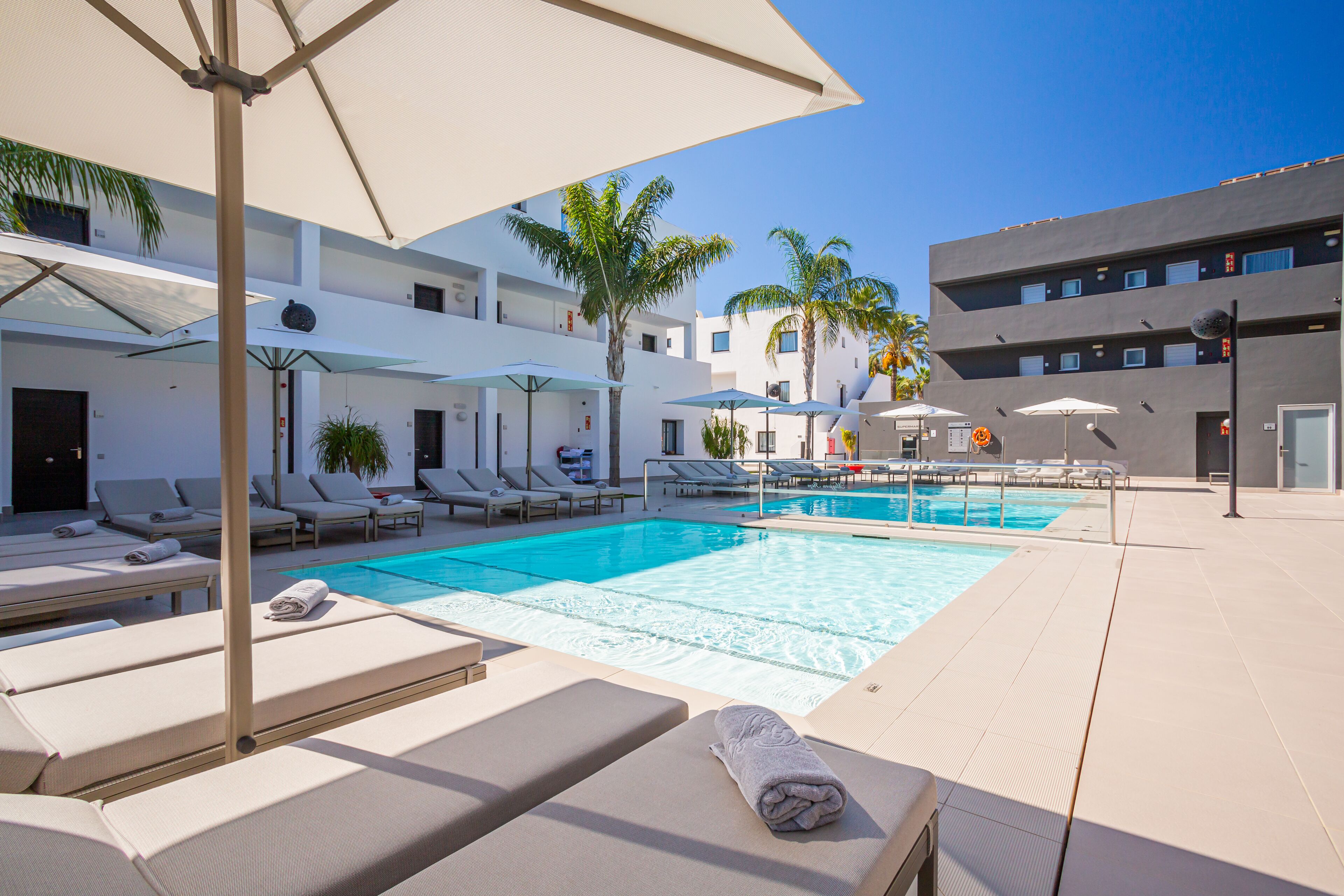 Un espace piscine aménagé avec des chaises longues et des parasols, une piscine à l’eau cristalline et des palmiers.