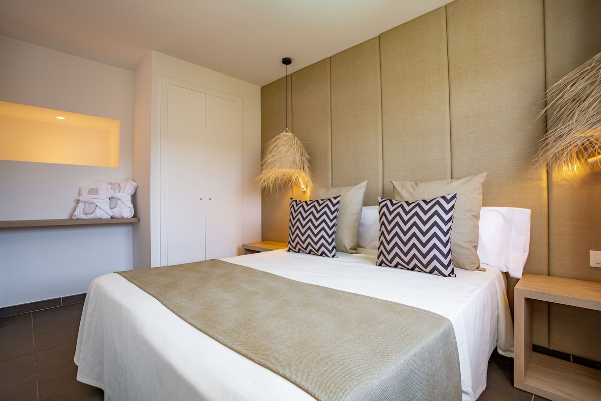 A modern hotel room with a large bed, a bedside table, a hanging light fixture, and a wall-mounted shelf with towels.