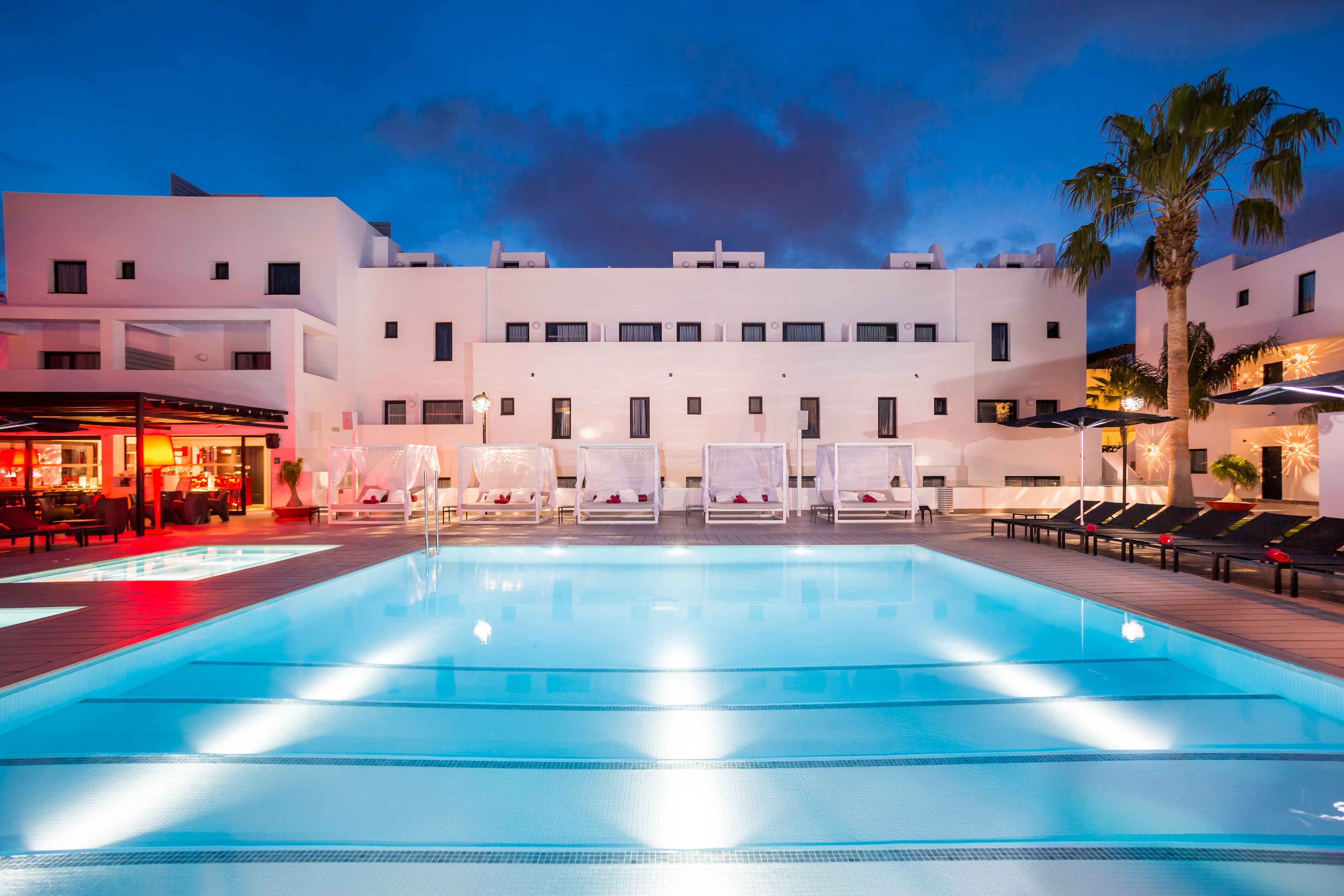 A swimming pool with lounge chairs and umbrellas, a bar area, and a building with multiple balconies.