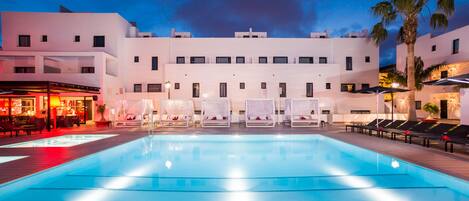 A swimming pool with lounge chairs and umbrellas, a bar area, and a building with multiple balconies.