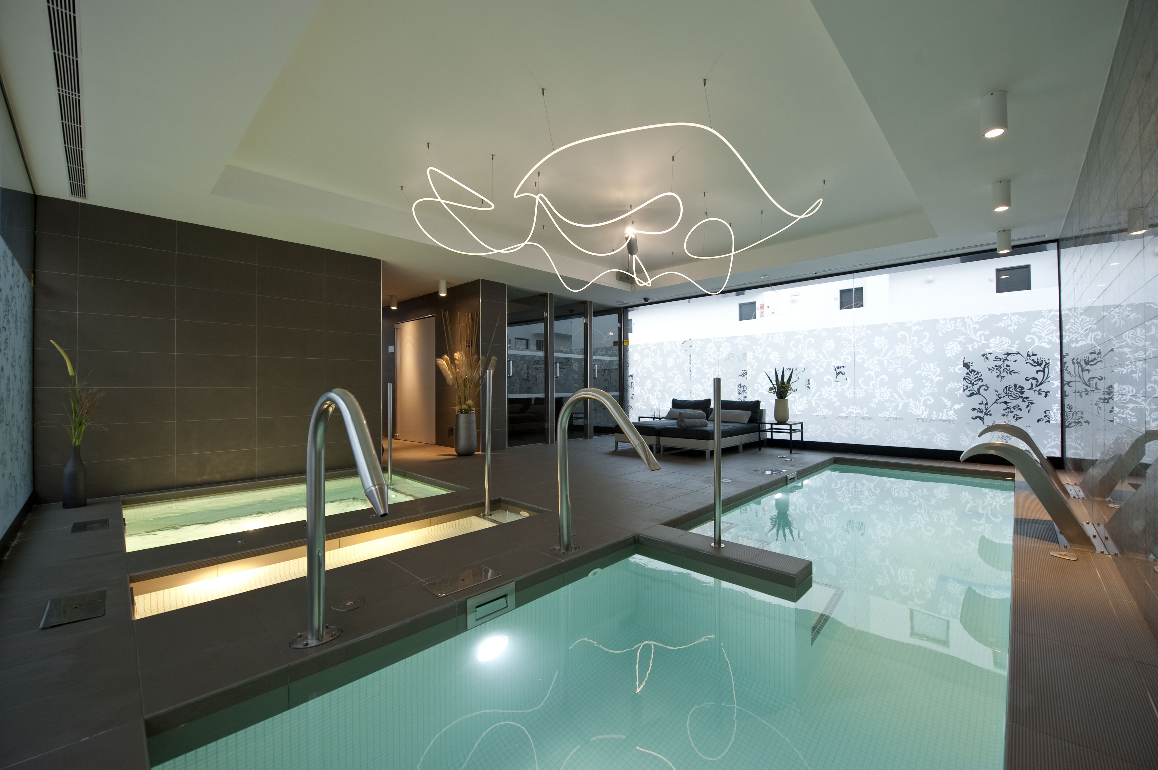 A modern indoor pool area with two separate pools, a lounge area with seating, and a decorative ceiling light.
