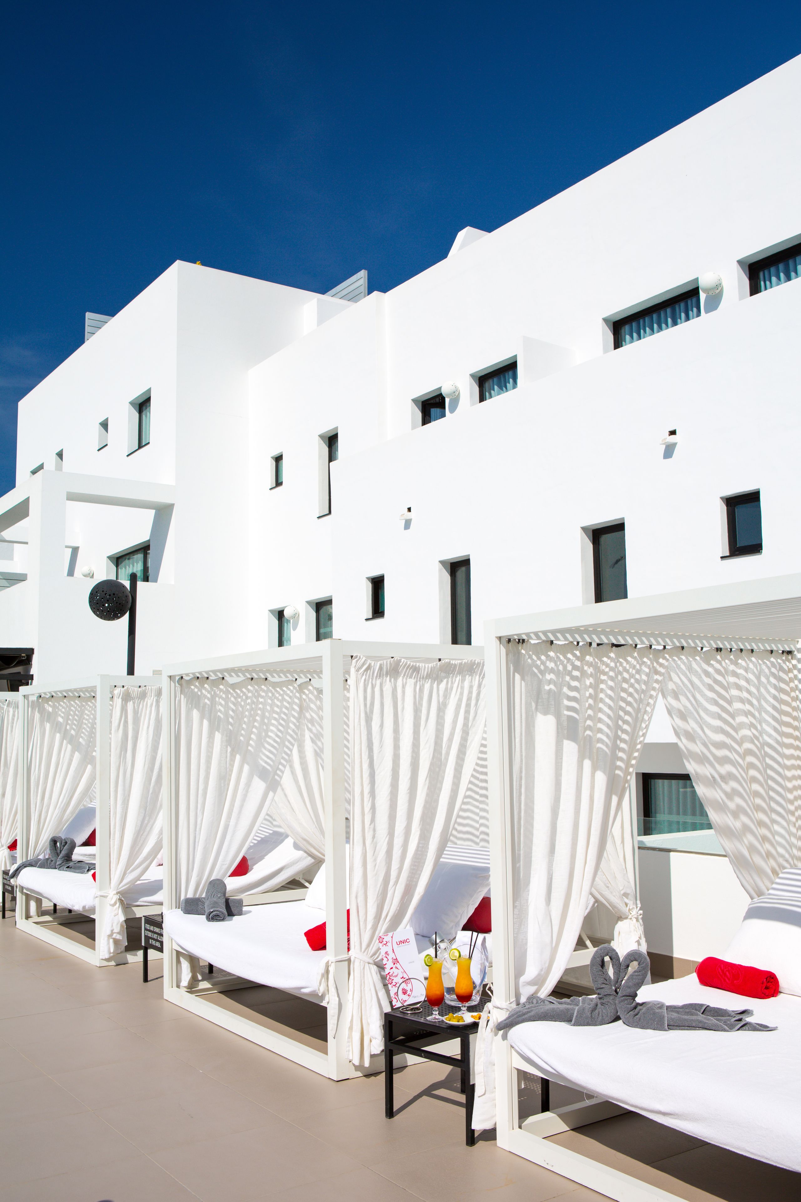 A row of white, single-room suites with canopy beds, each featuring a small table with a drink and a towel.