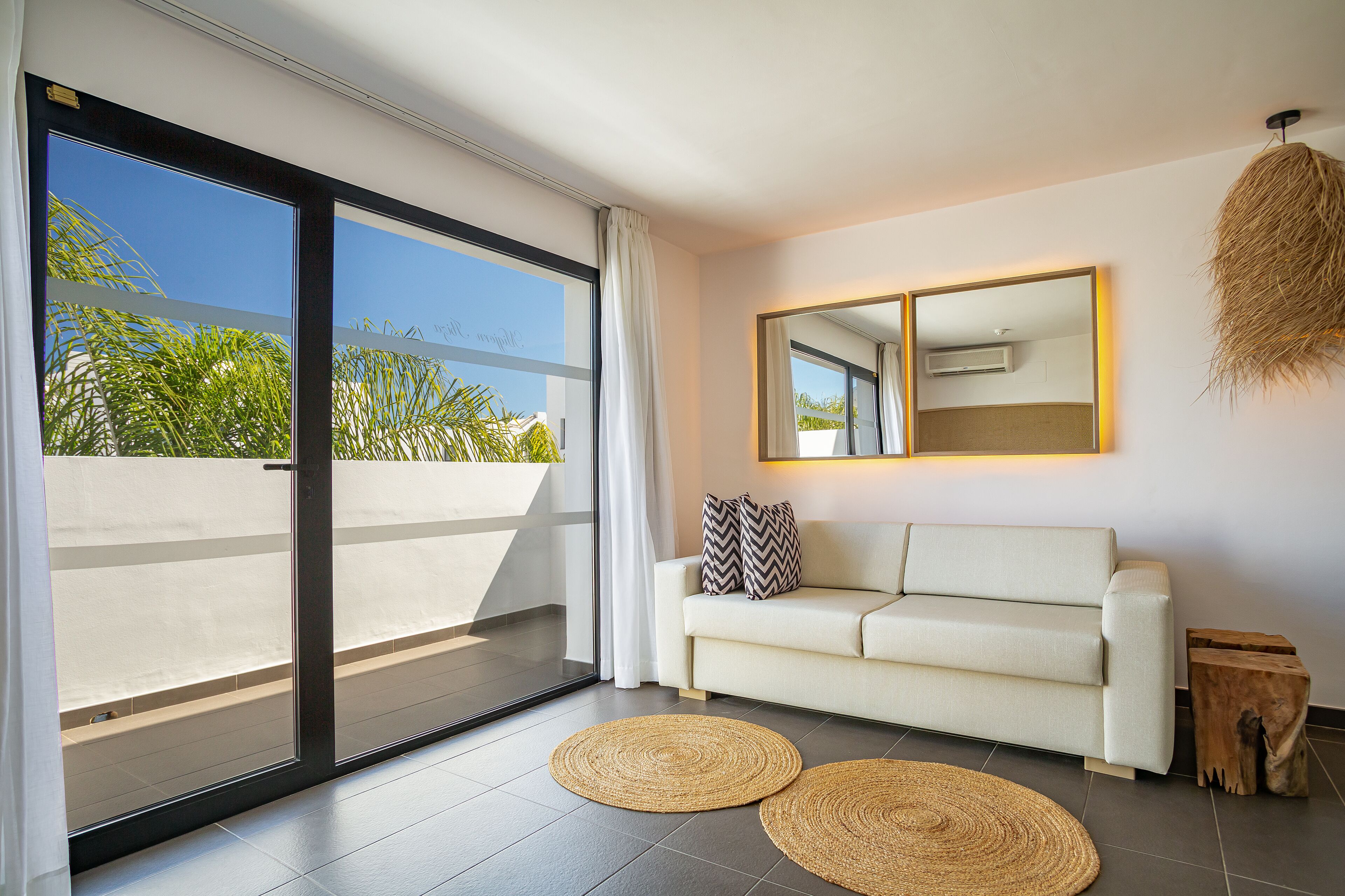 A modern living room with a white sofa, two woven rugs, a large sliding glass door, and a mirror on the wall.