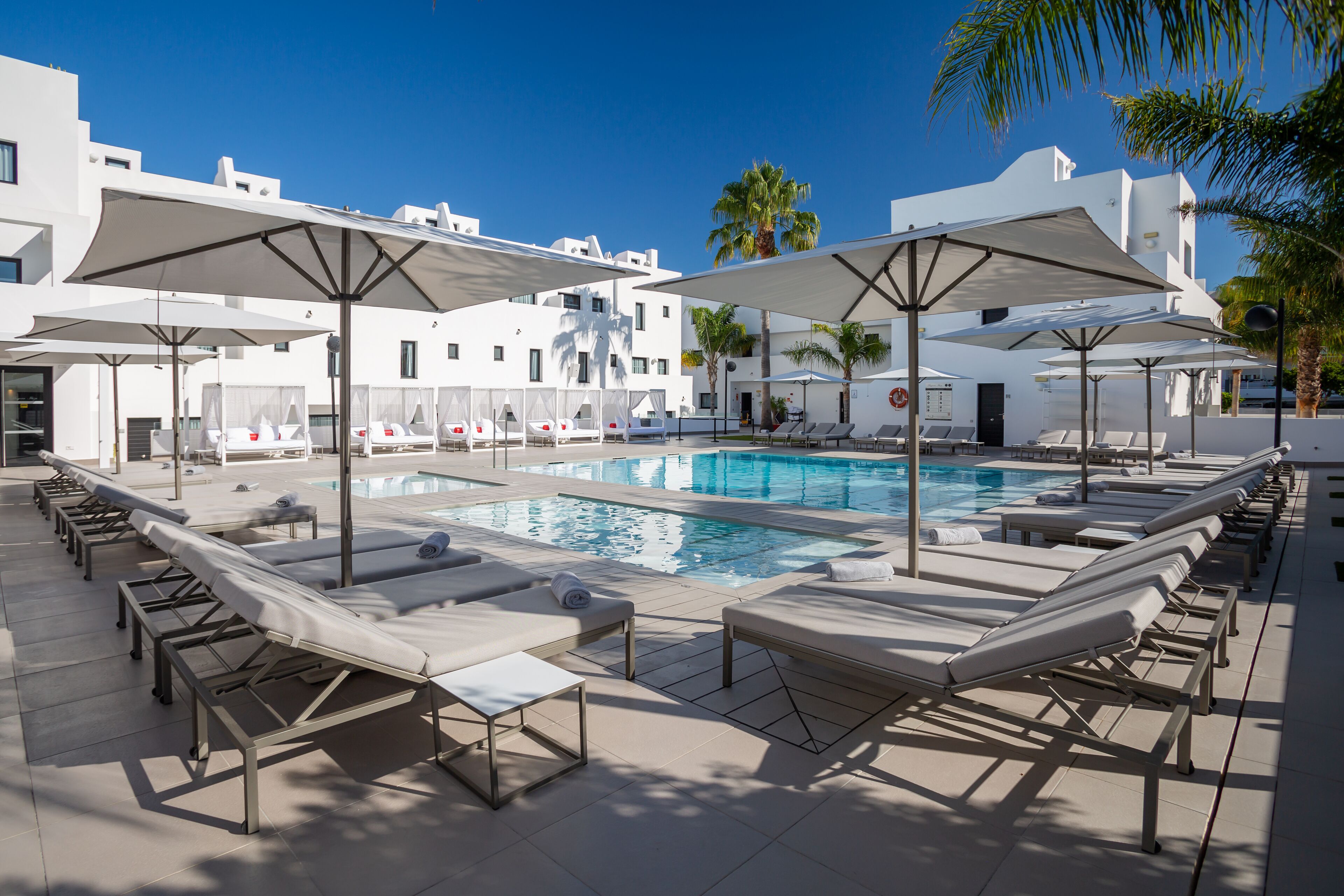 Un espace aménagé au bord de la piscine, avec des chaises longues et des parasols, un ciel d’un bleu éclatant et des bâtiments blancs en arrière-plan.