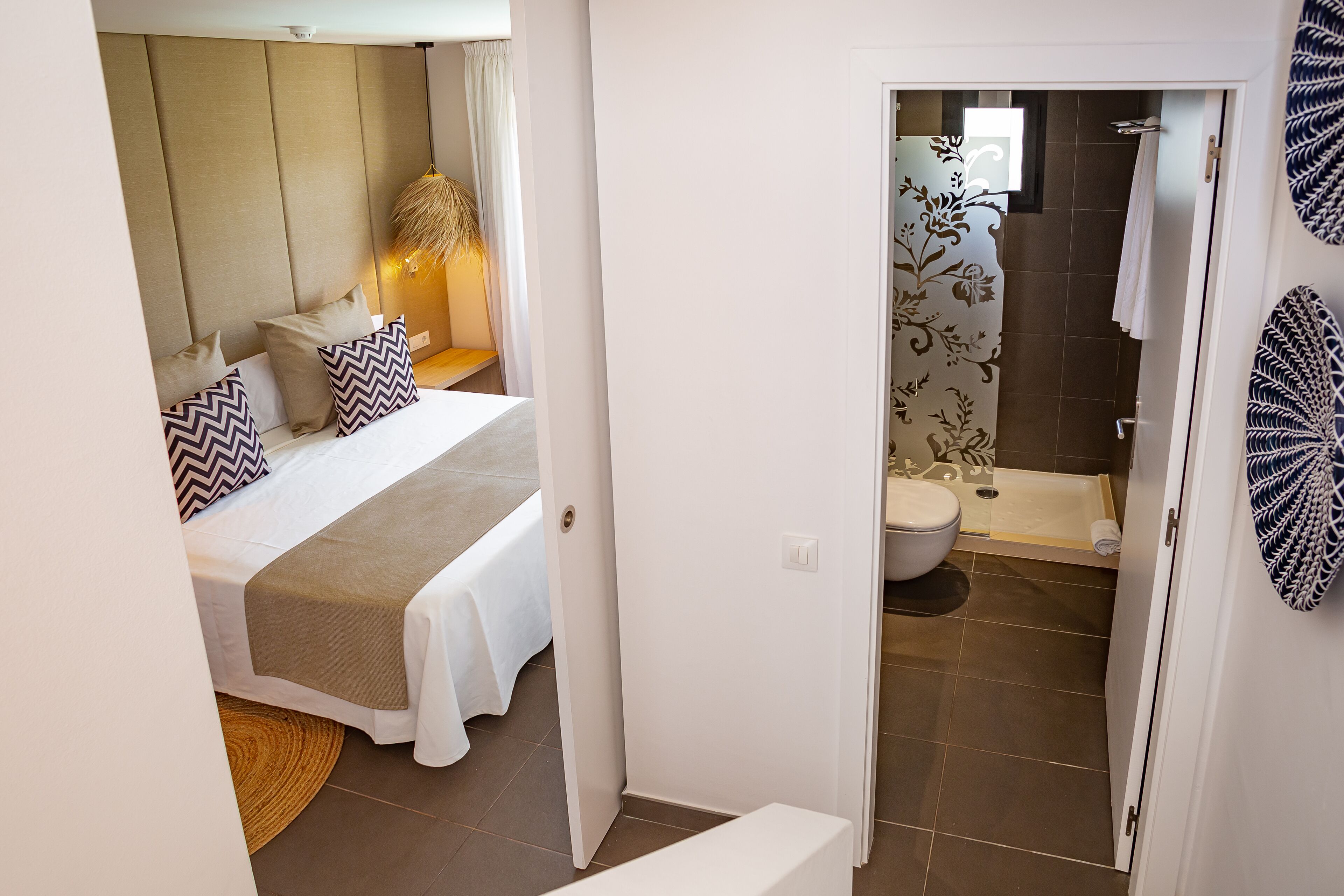 A bedroom with a bed, bedside table, and a lamp. There is a bathroom with a toilet, shower, and a towel rack.