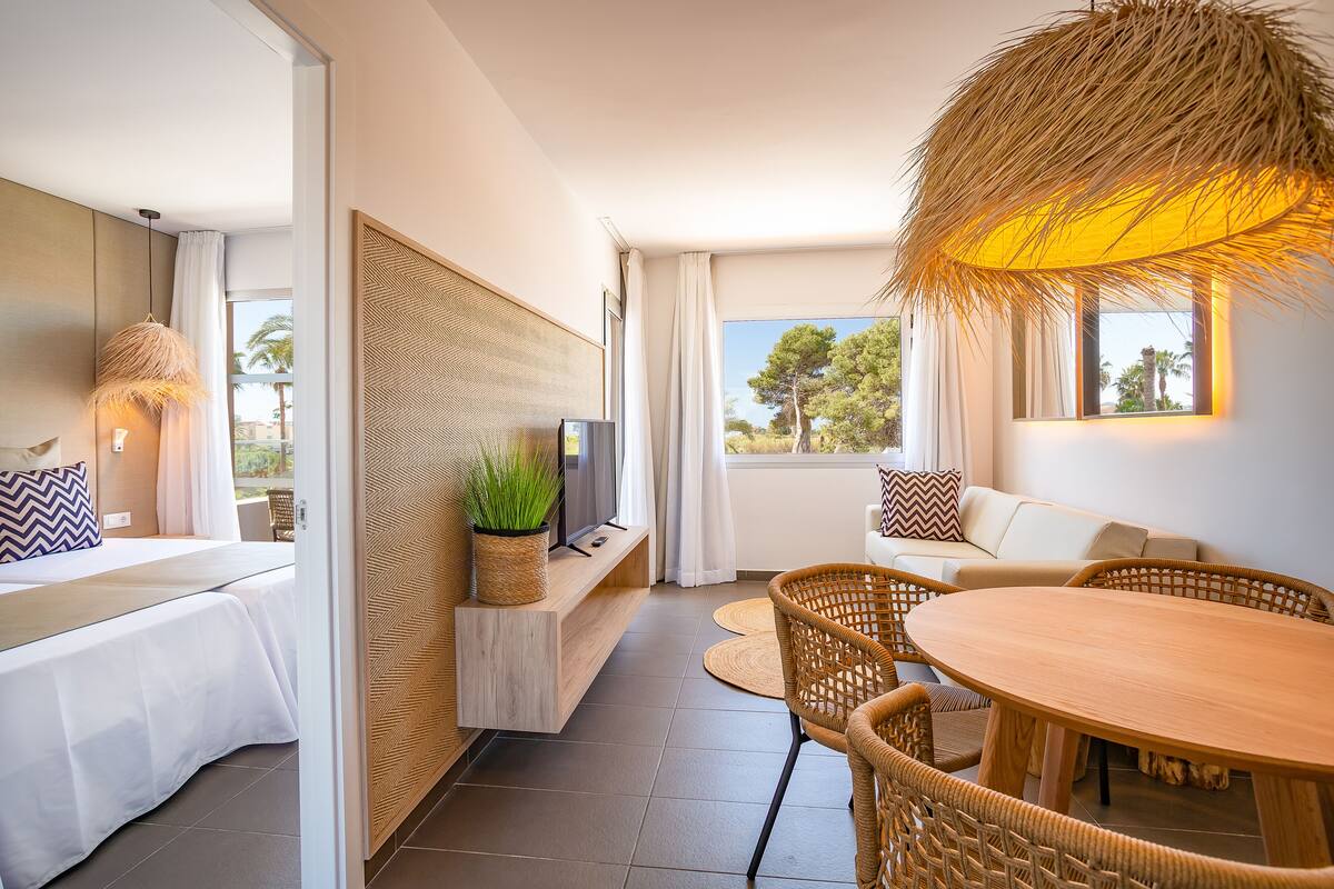 A modern hotel room with a bed, a sofa, a dining table, and a TV.