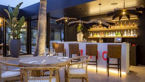 A modern bar area with wicker furniture, a large potted plant, and a variety of bottles on shelves. - Migjorn Ibiza Suites & Spa (Sant Josep de sa Talaia)