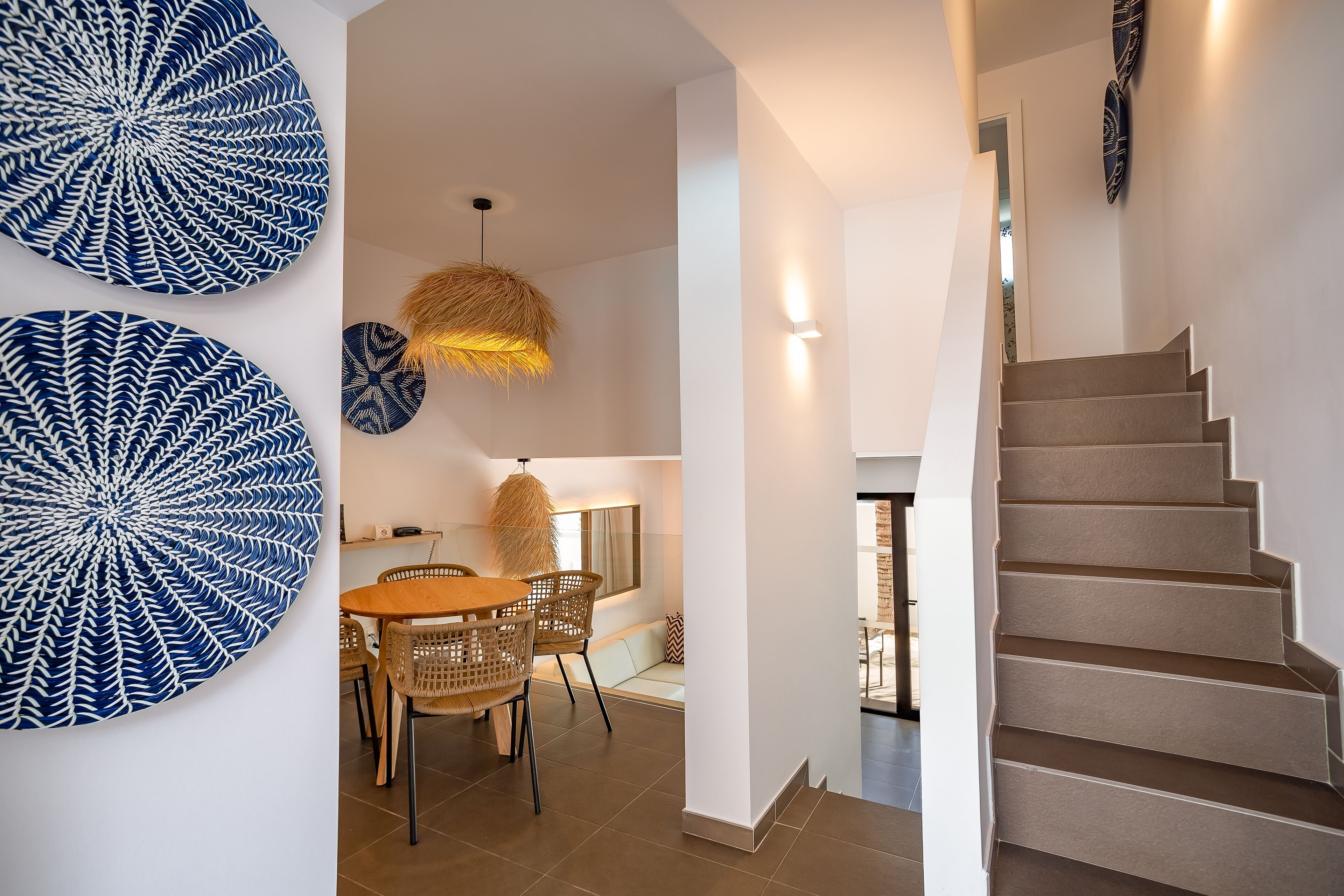 Een modern interieur met een trap, een eethoek met rieten stoelen en decoratieve muurkunst.