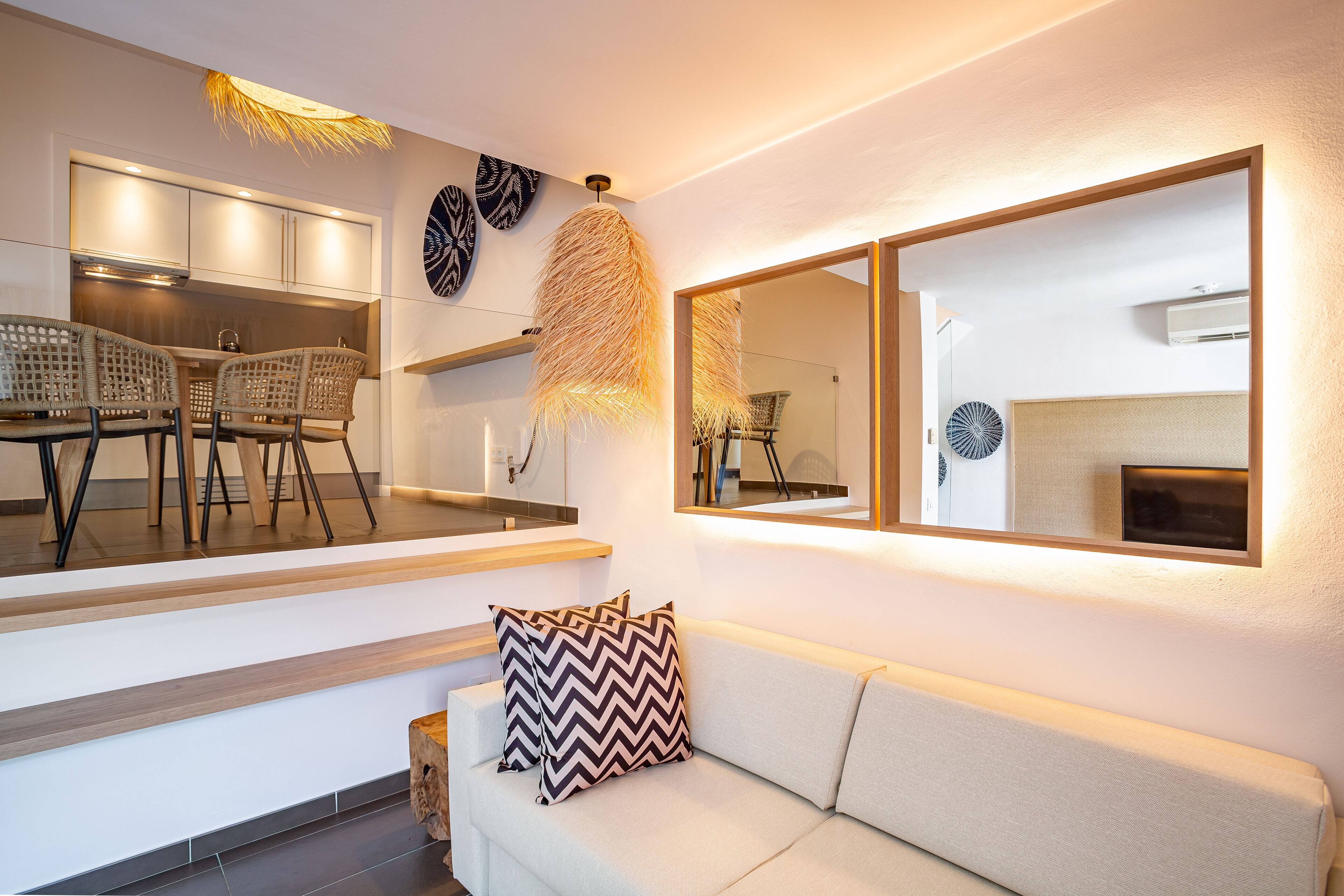A modern living room with a sofa, a dining area with chairs, a kitchen with cabinets, and a large mirror on the wall.