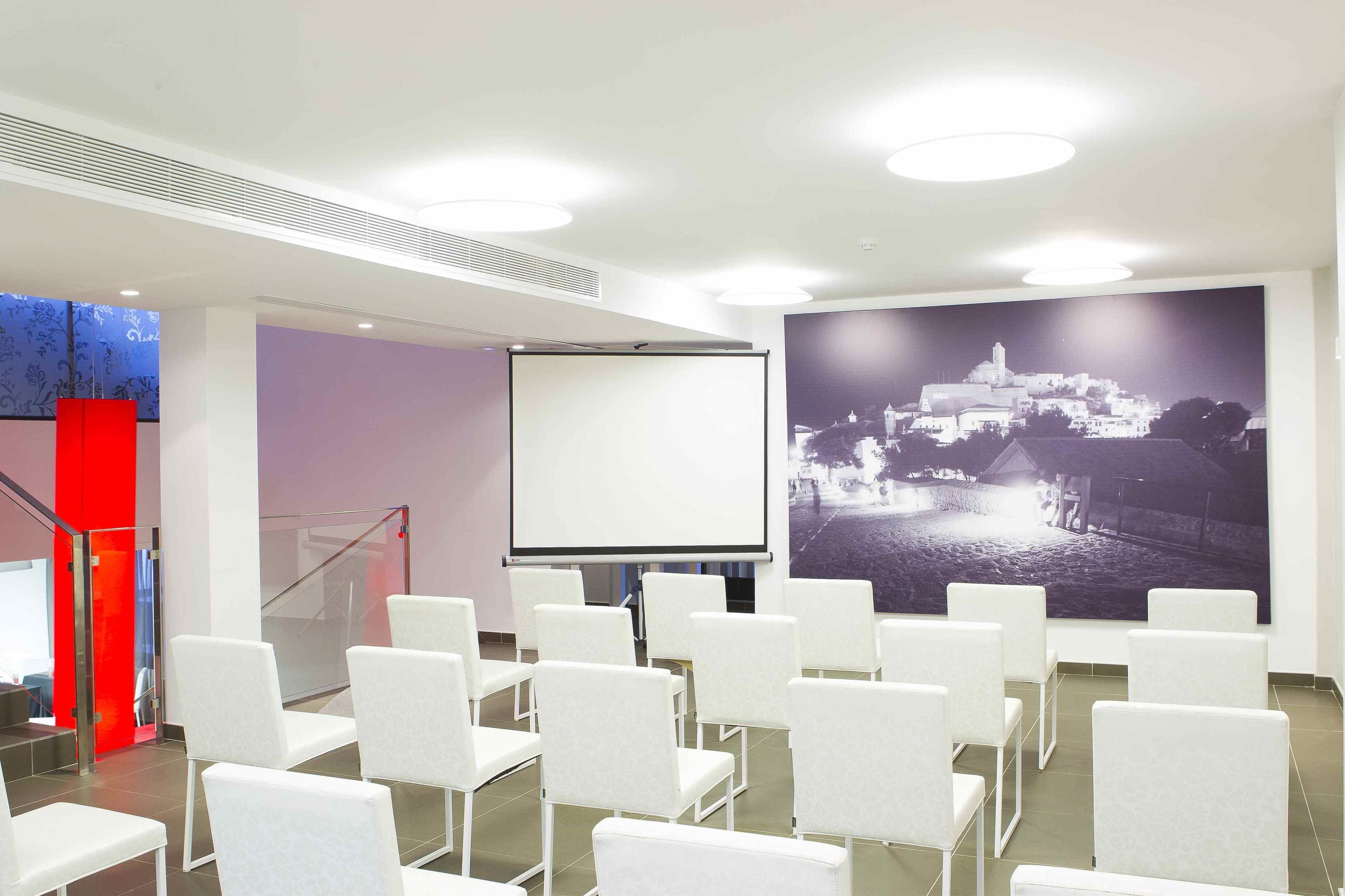 A conference room with white chairs arranged in rows, a projector screen, and a large black-and-white photograph on the wall.