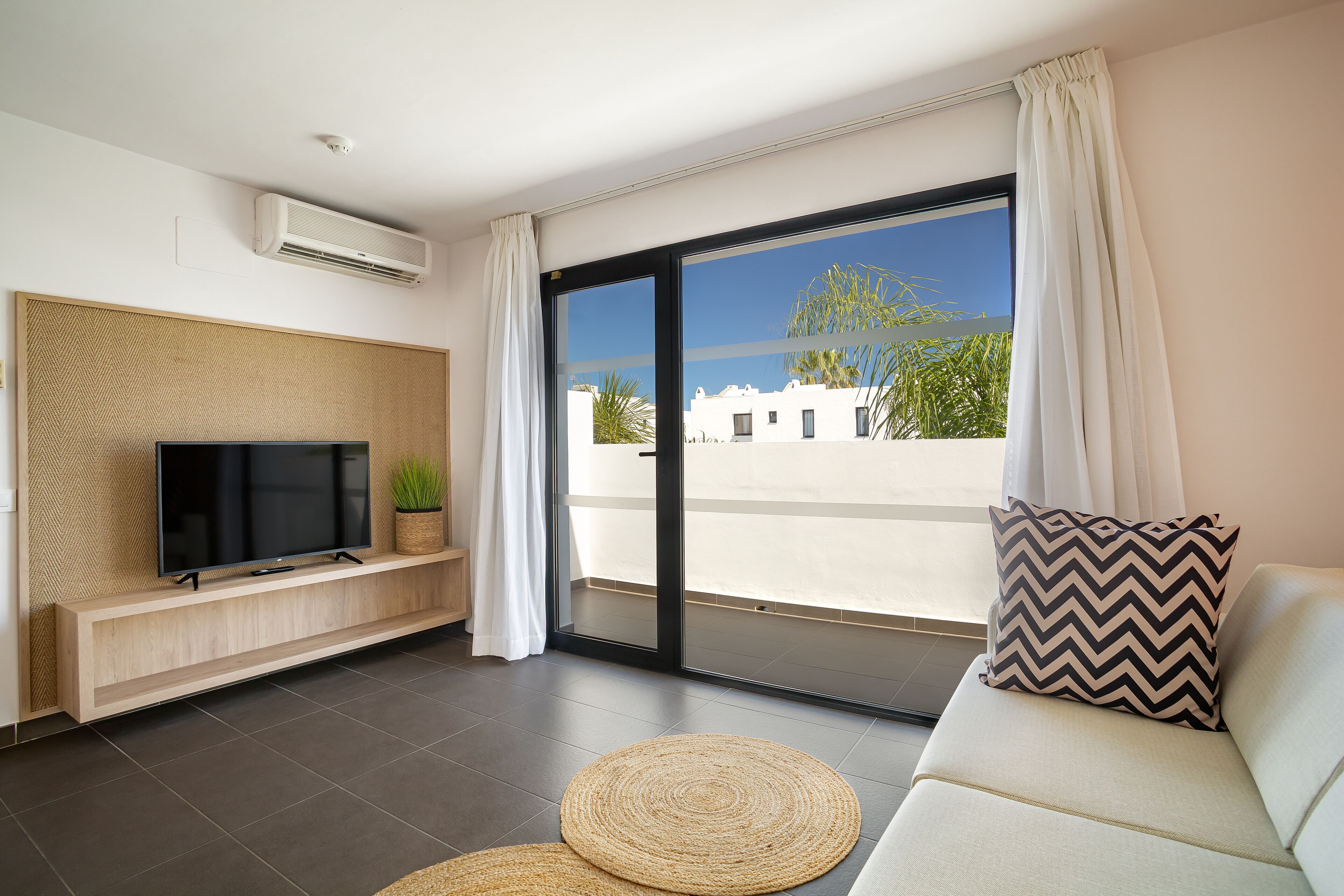 A modern living room with a flat-screen TV on a wooden console, a sofa with a patterned pillow, and a view of a white building through large glass doors.