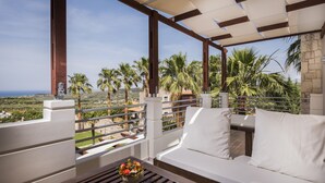 A balcony with a white sofa, a wooden table with a flower vase, and a view of palm trees and the sea. - Athina Luxury Villas (Platanias)