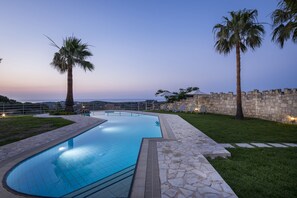 Una piscina con baldosas de piedra, rodeada de césped y palmeras, con un muro de piedra y sillones de descanso al fondo.