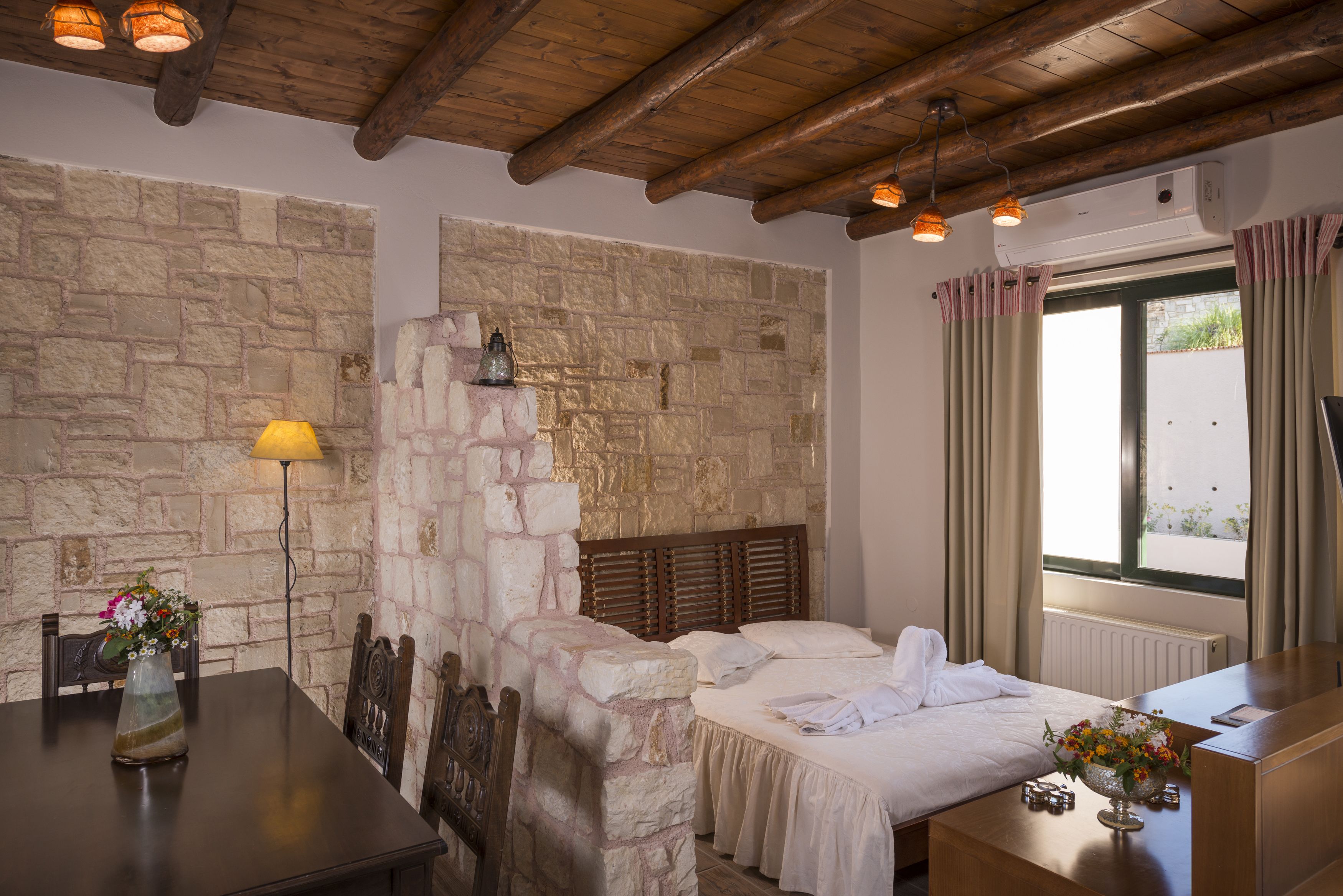 A room with a stone wall, wooden ceiling beams, a bed with white linens, a bedside table with a vase of flowers, and a window with curtains.