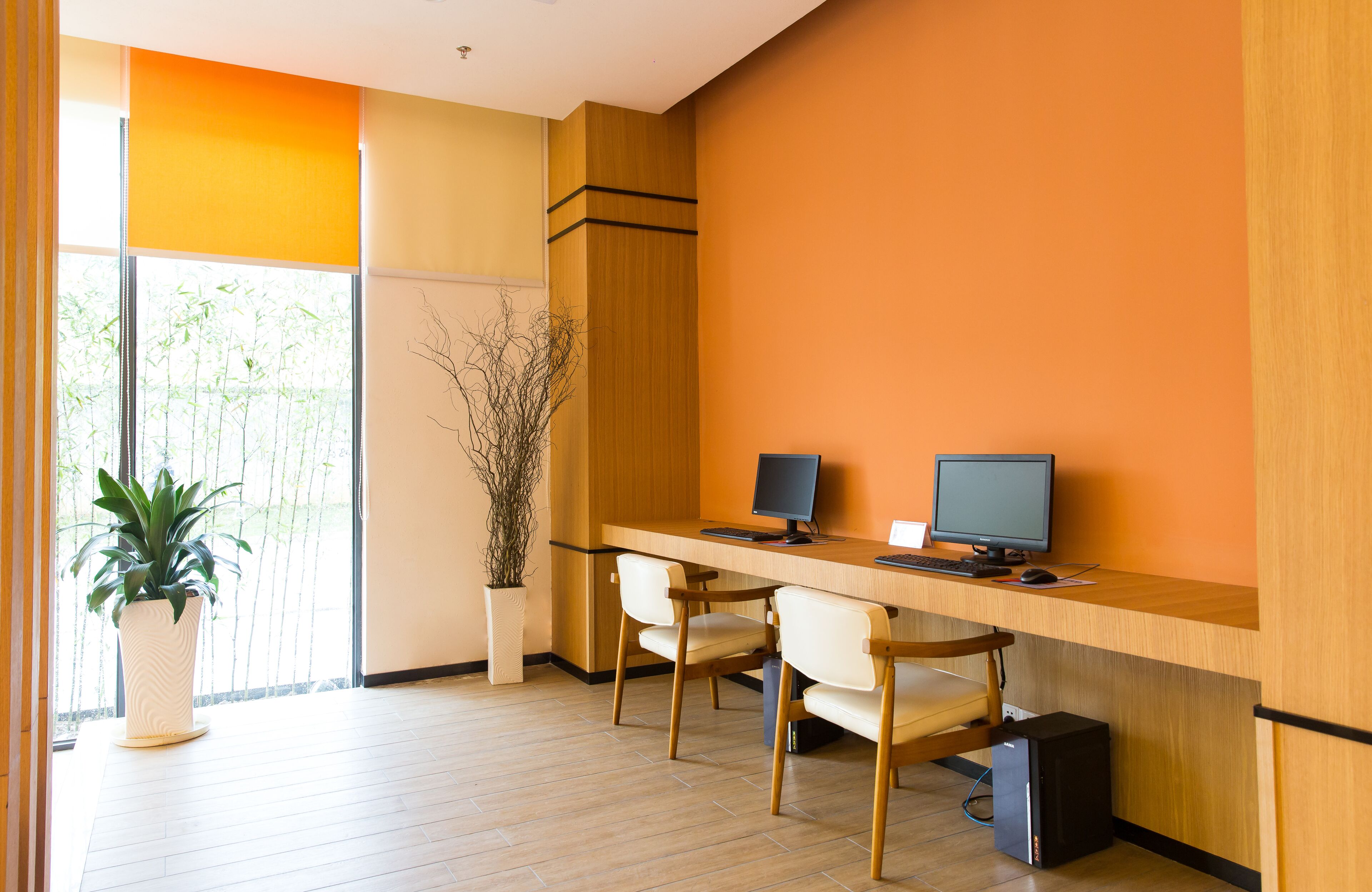A modern office space with a long wooden desk, two computers, two chairs, a potted plant, and a large window with a view of trees.