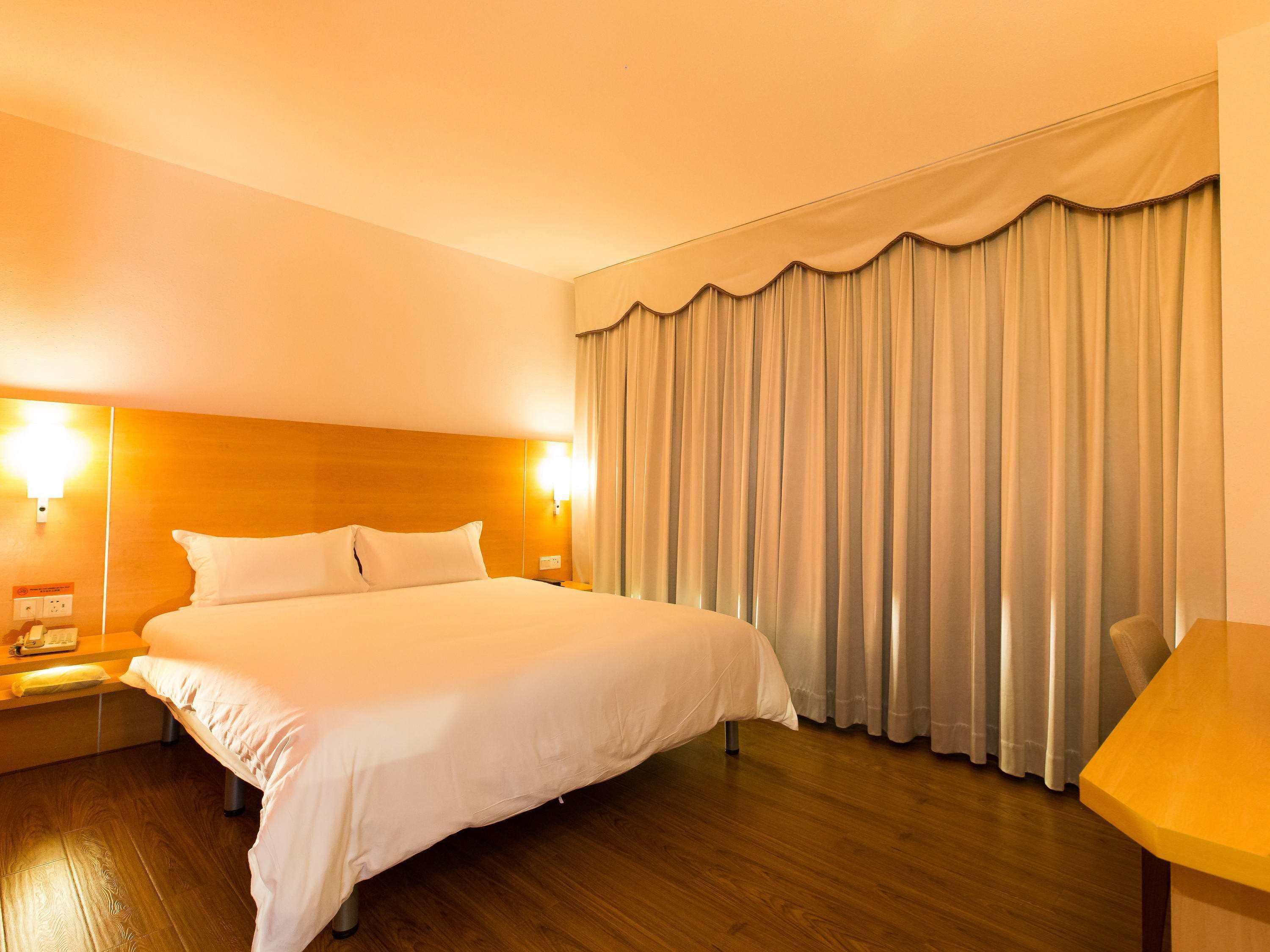 A hotel room with a large bed, wooden flooring, a nightstand with a phone, and a desk with a chair.