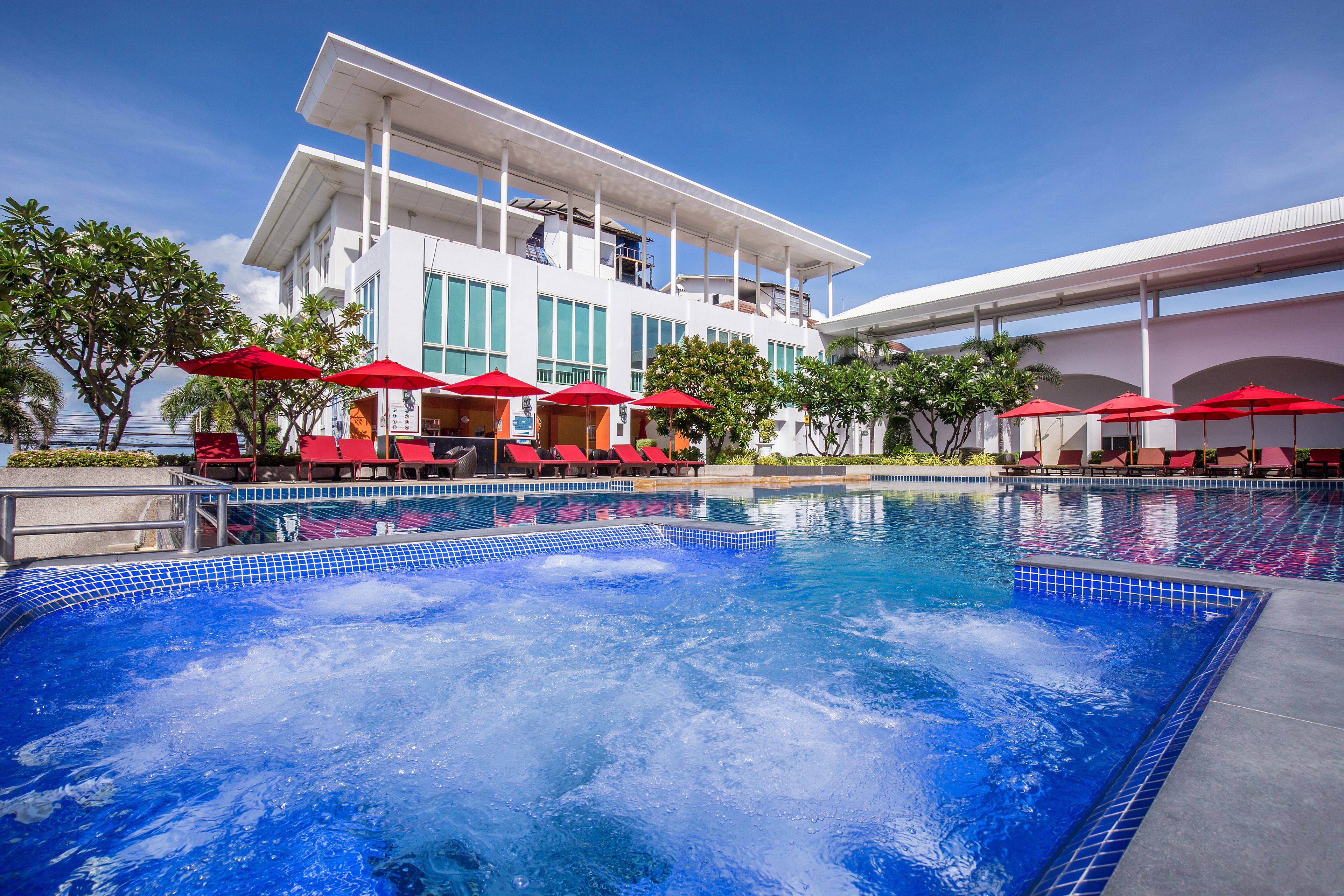 A swimming pool with red umbrellas and lounge chairs, surrounded by a modern building with large windows and a covered walkway.