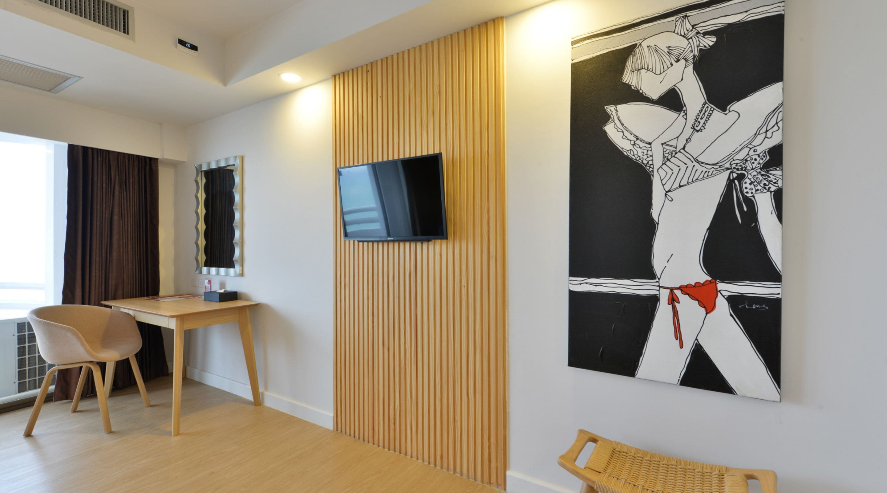 A modern hotel room with a wooden panel wall, a flat-screen TV mounted above a desk, a large abstract painting, and a wicker chair.