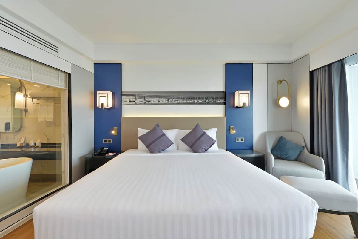 Une chambre d’hôtel moderne dotée d’un grand lit, d’un coin salon avec un fauteuil et d’une salle de bain séparée par une paroi en verre.